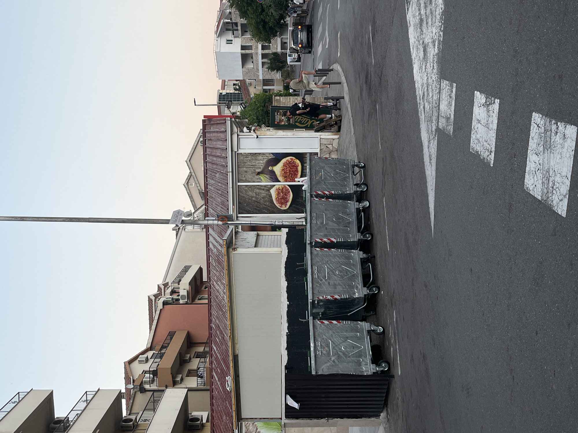 In Budva, Montenegro, a street scene is captured at dusk. In the foreground, a paved road features white road markings, including a pedestrian crossing. Several large, silver-colored trash bins with wheels are lined up on the left side of the road. Behind the trash bins is a building with a corrugated metal roof and a beige facade. A large advertisement for figs is displayed on a window of the building.
Further down the street, a few people are visible. Two individuals are standing near a black scooter, while another person is leaning on a railing. A car is driving on the road, and several residential buildings with balconies are seen in the background. The sky is a clear, pale blue, indicating the end of the day. The overall impression is a typical urban streetscape with a commercial element.
