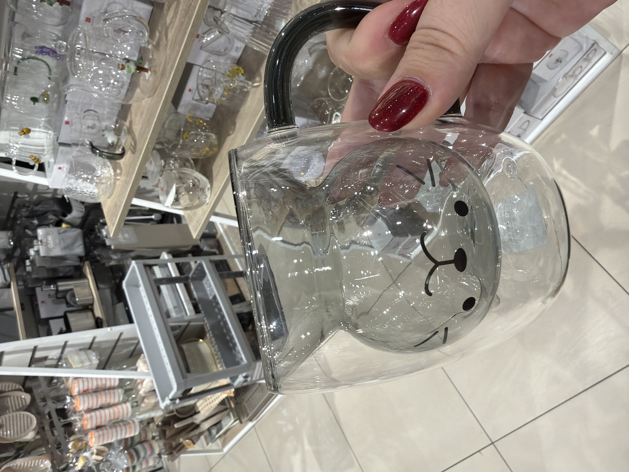 A close-up shot captures a clear glass pitcher with a black handle, bearing a simple cartoon-like face of an animal, possibly a bear or cat, featuring two eyes, a nose, and whiskers. The pitcher is held by a hand with burgundy nail polish. The background consists of shelves filled with various items, including glassware, kitchenware, and other household products. The items are displayed on wooden shelves, suggesting a retail environment, likely a store in the city of Kazan, Russia. The lighting is bright and artificial, typical of a retail space.