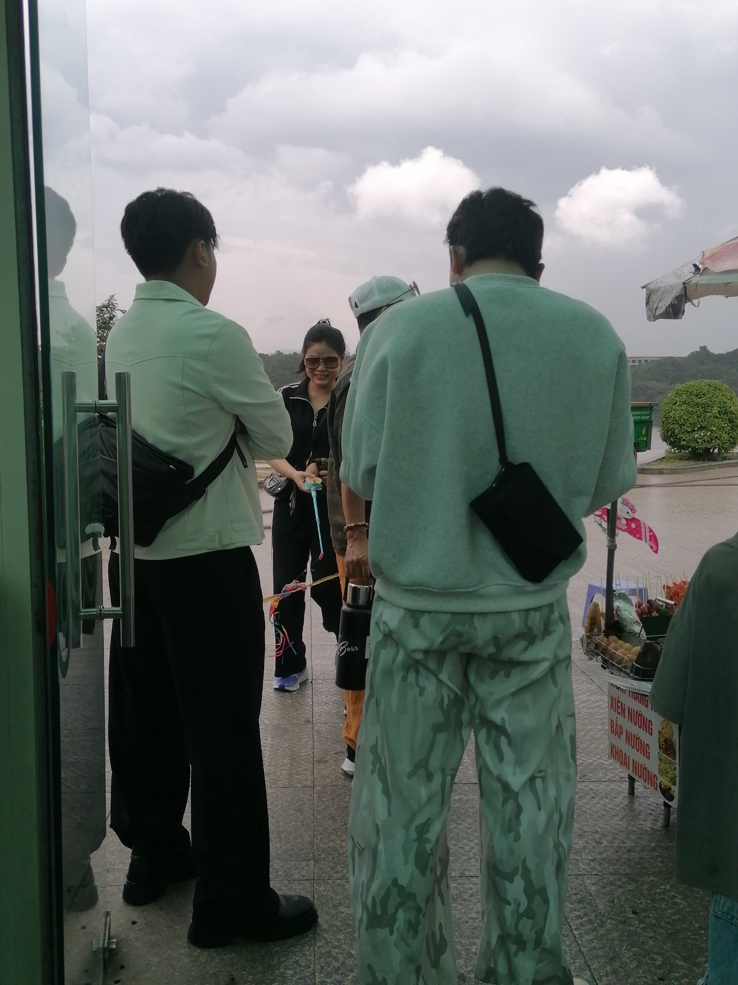 The image depicts a scene at a street food stall in Da Lat, Vietnam, under an overcast sky. Two men and a woman are the primary subjects. A man in a light-colored jacket and black pants stands facing left, with a black crossbody bag. A woman in black athletic wear with sunglasses and her hair in a bun is interacting with him. Behind them, another man in a light green sweatshirt and camouflage pants faces away from the camera, also with a black crossbody bag. A street food stall is visible to the right, displaying skewers of food and some packaged items. The ground is paved with square tiles. The environment suggests an outdoor, possibly tourist-oriented area.