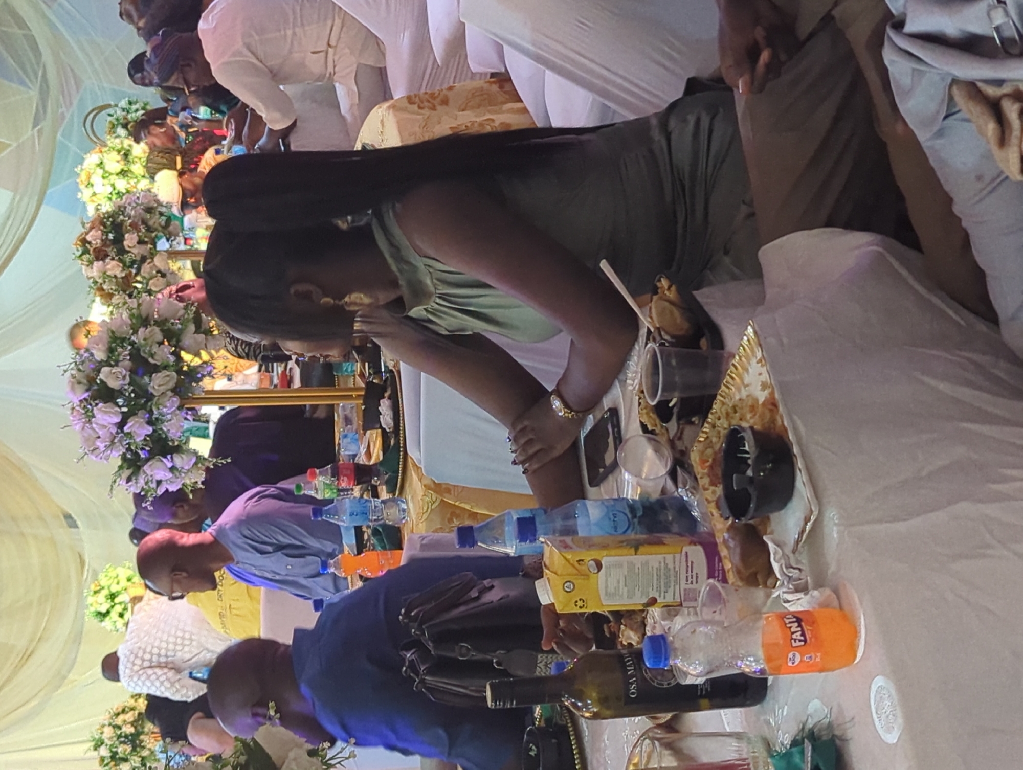 This image depicts a celebratory gathering, possibly a wedding or a formal event, taking place indoors in Barden Yana, Nigeria. The scene is set around tables laden with food and drinks. In the foreground, a woman in a green sleeveless dress with a high neckline is seated, her back mostly towards the viewer. She has long black hair styled in a high ponytail and is wearing large gold earrings. Her left arm is extended across the table, resting on some items. To her left, a bottle of Osa Mayi wine and a carton of juice are visible. A bottle of Fanta is also prominent on the table.
Other individuals are visible in the background, seated at various tables. One man in a blue shirt with a pattern is looking to his right. Another person in a white outfit with intricate patterns is partially visible in the upper left quadrant. Floral arrangements, predominantly white and pink, adorn the space, suggesting a festive atmosphere. Drapes of fabric in shades of cream and yellow form a canopy overhead, contributing to the formal ambiance. The lighting appears to be a mix of ambient and possibly artificial light, casting soft shadows. The overall impression is that of a well-attended event with a focus on dining and socializing.