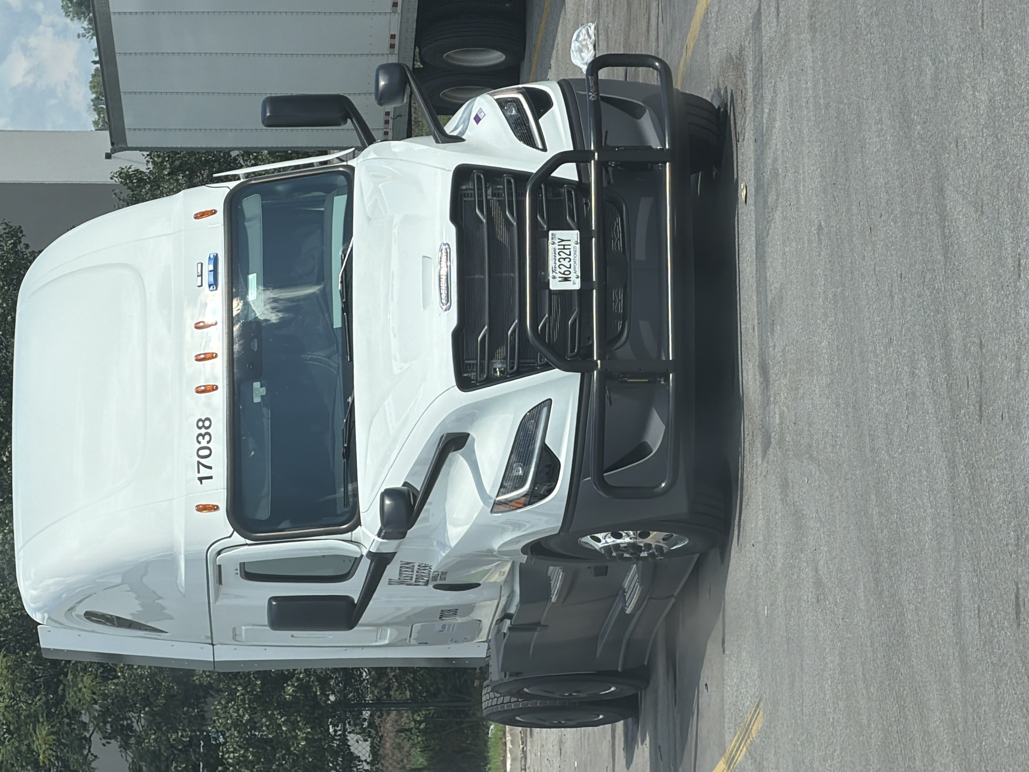 A white Freightliner semi-truck with the company logo "WESTERN EXPRESS" is parked in an asphalt lot. The truck has a license plate that reads "W6232HY" and is from Tennessee. A second semi-trailer is visible behind the truck, on the right side of the image. The scene appears to be in a commercial area, possibly a truck stop or distribution center, based on the presence of multiple semi-trucks and a fenced area with trees in the background. The sky is partly cloudy.