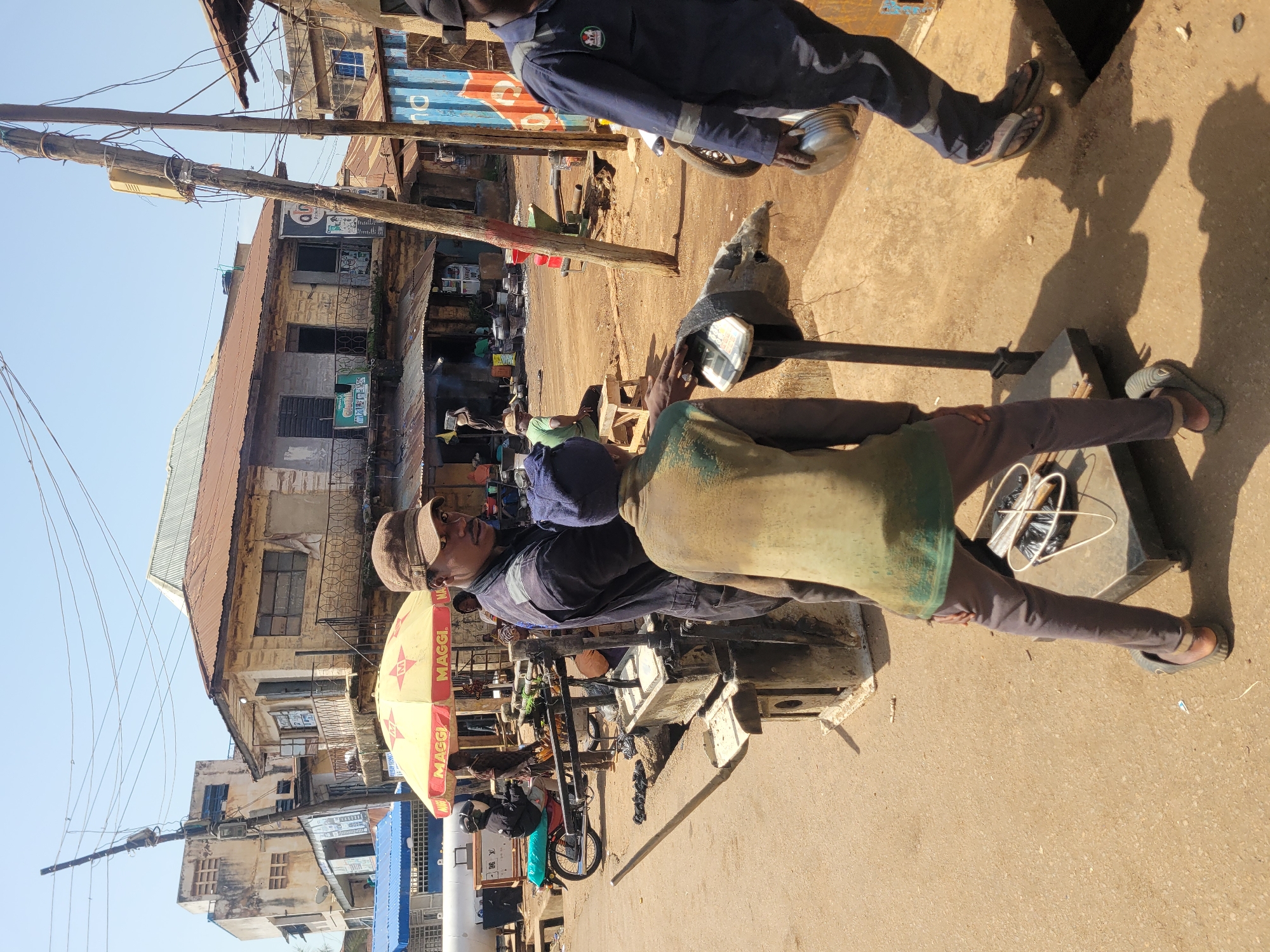 The image depicts a street scene in Jos, Nigeria, likely a market area. In the foreground, two individuals are engaged with a weighing scale. One person, wearing a dark blue jumpsuit and a cap, stands on the right, holding a metal object, possibly a container, in their right hand. They appear to be a worker, indicated by their attire. The second individual, facing away from the camera and dressed in a green shirt and brown pants, is interacting with the scale, which is covered with a worn grey fabric.
The background reveals a bustling environment with several buildings, some appearing aged and in disrepair, with corrugated iron roofs. A yellow umbrella with "MAGGI" branding is visible, suggesting commercial activity. A motorcycle with a rider wearing a helmet is parked to the left, partially obscured by the weighing scale setup. Various other individuals and market stalls can be seen further in the background, hinting at a busy marketplace. Electrical poles with numerous wires crisscross overhead. The ground is unpaved and dusty, typical of many street scenes in the region. The lighting indicates a bright, sunny day.