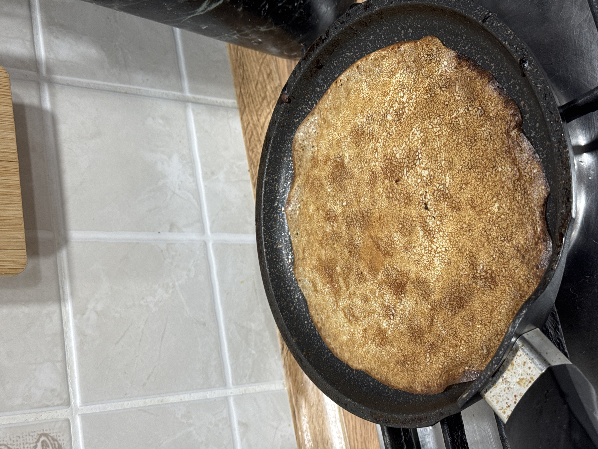 A pancake is being cooked in a black frying pan on a stovetop. The pancake is golden brown with a textured surface. The pan is on the right side of the image. To the left is a tiled counter. The city and location are unknown.