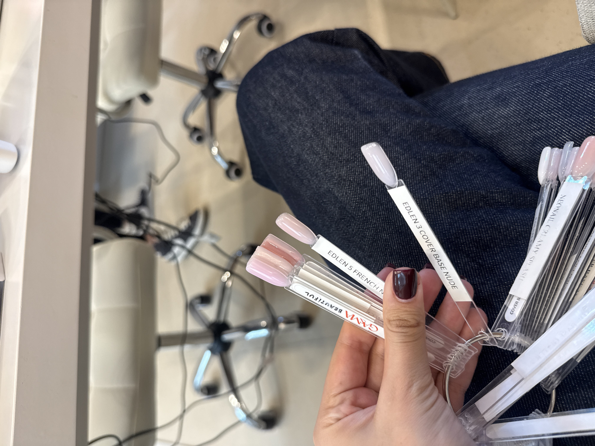 In an indoor setting, likely a nail salon or beauty studio in Milan, Italy, a person is shown from a first-person perspective, seated with their legs, clad in dark blue jeans, visible in the frame. Their left hand, adorned with dark red nail polish on the thumb, is holding several rings of nail polish swatches. The swatches display various nude, pink, and light-colored polishes, suggesting the person is in the process of selecting a shade.
Visible text on the swatches includes brands and specific colors: "EDLEN 3 COVER BASE NUDE," "EDLEN 5 FRENCH N," and "GAMA BEAUTIFUL." Partially visible text includes "NEONAIL CREAMY SPLASH," "COVER B," and "LER RASE." In the background, blurry elements of white salon chairs with chrome wheeled bases are visible, along with parts of a white table or counter on the left, reinforcing the salon environment. The scene captures a moment of aesthetic decision-making.