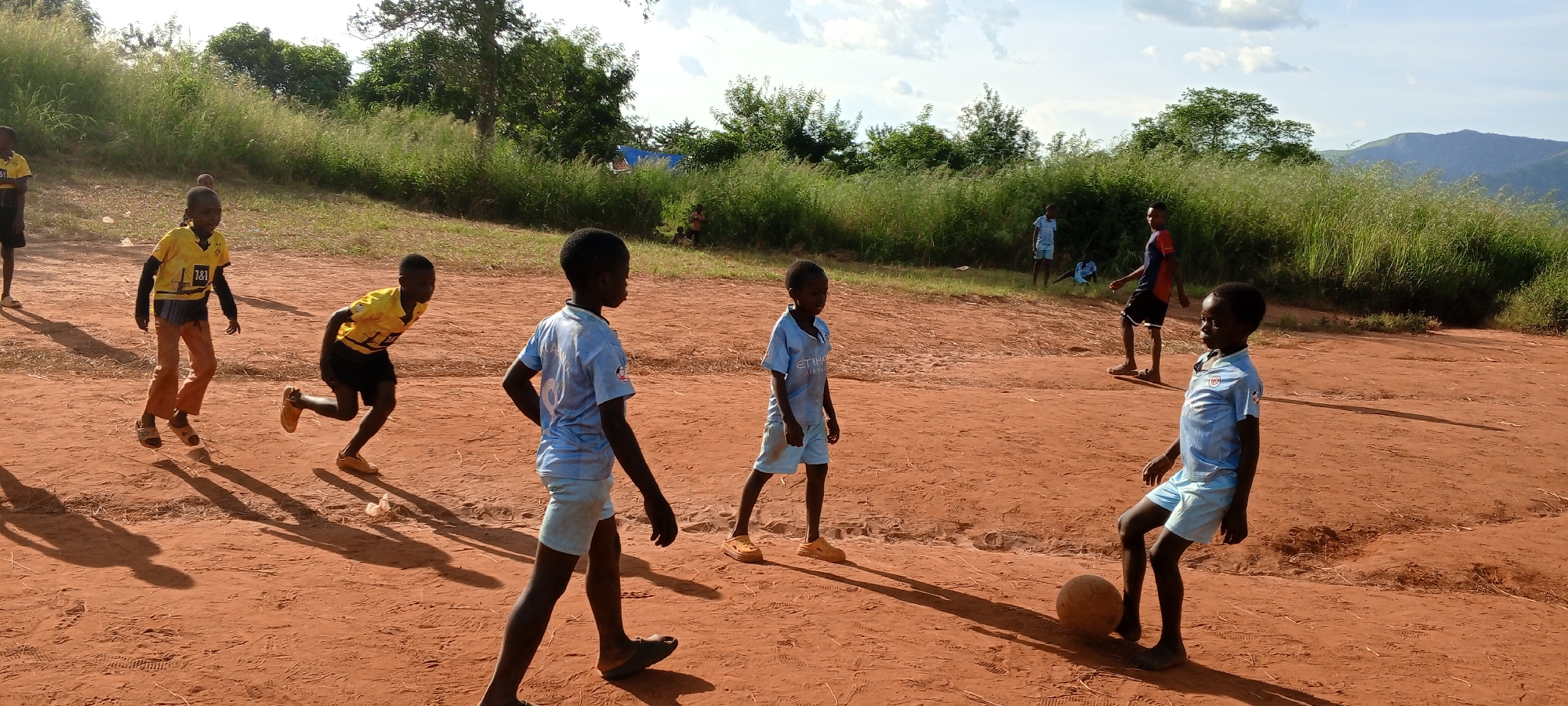 In an outdoor area with a reddish-brown dirt surface in Ngalula, Democratic Republic of the Congo, a group of boys are engaged in a soccer game. Some boys wear light blue soccer uniforms while others wear yellow soccer jerseys. Some of the boys are running. One boy stands by a soccer ball. A grassy hillside is behind the boys. Trees and mountains are visible in the background.