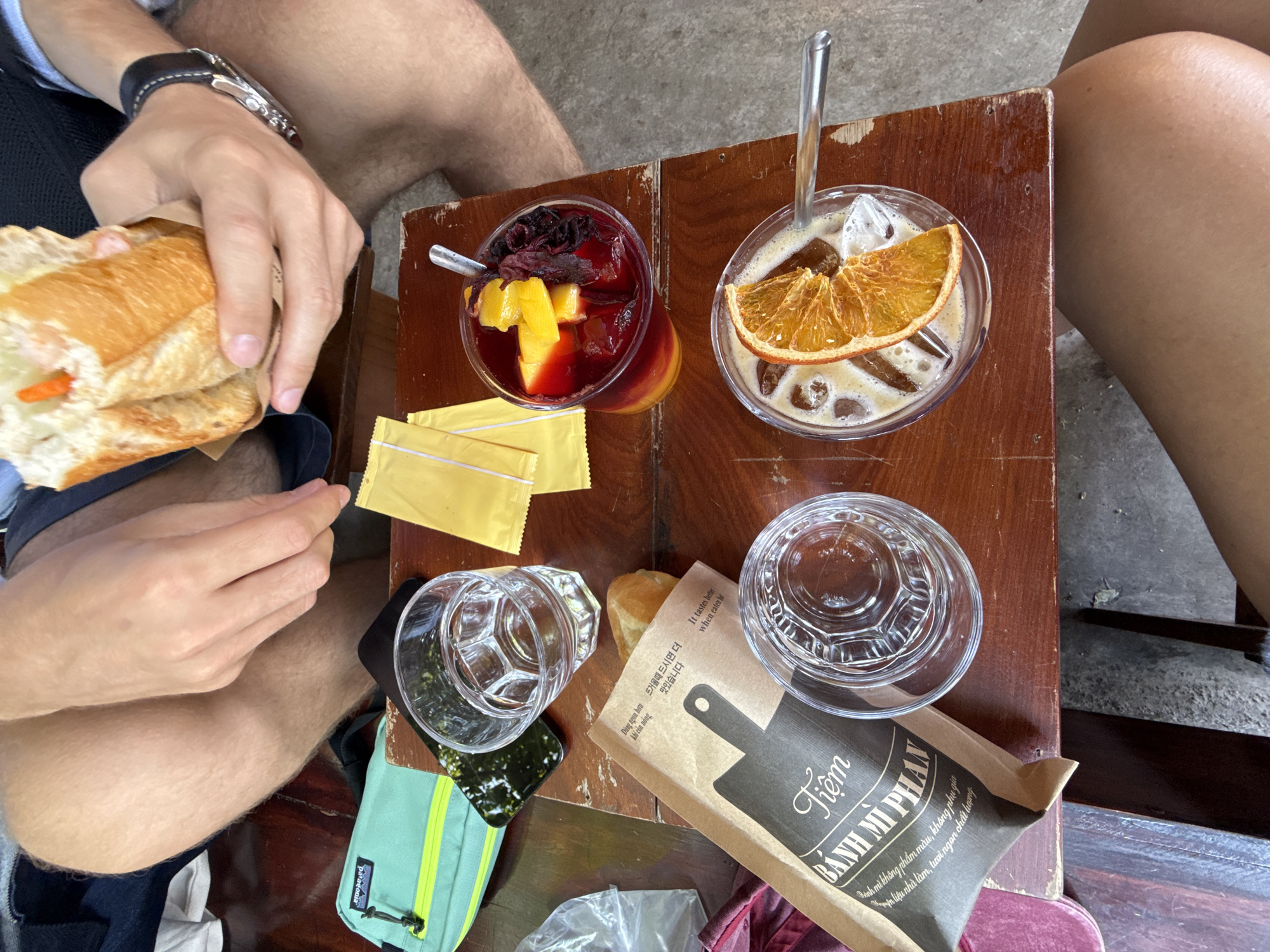 An overhead shot captures two individuals enjoying a meal and drinks at a wooden table, likely outdoors. One person's hands are visible, holding a Banh Mi sandwich wrapped in paper, suggesting they are about to eat or have just started. A watch is visible on this person's wrist. The other person's legs are visible to the right.
The table holds several items:
- A red-colored drink in a clear glass, garnished with what appears to be dried hibiscus petals and pieces of mango or similar yellow fruit. A spoon is in the glass.
- An iced coffee drink in another clear glass, topped with a dried orange slice and featuring a spoon.
- Two empty clear glasses.
- Yellow packets, possibly sugar or condiments.
- A small mint green pouch with a yellow zipper and strap, bearing the "Patagonia" brand name.
- A brown paper bag or wrapper, prominent in the foreground.
The paper packaging features the text "Tiệm BÁNH MÌ PHAN" (Banh Mi Phan Shop) with additional text in Vietnamese, Korean, and English. The Vietnamese text reads: "Bánh mì không phẩm màu, không phụ gia nguyên liệu nhà làm, tươi ngon chất lượng" (Banh mi without artificial colors, no additives, homemade ingredients, fresh and quality) and "Dùng ngon hơn khi còn nóng" (Tastes better when hot). The English translation "It tastes better when eaten hot" is also present, alongside the Korean "맛있습니다" (Delicious).
The setting suggests a casual eatery or street food stall, common in places like Nha Trang, Vietnam, where Banh Mi is a popular staple. The presence of iced drinks indicates warm weather, likely daytime. The table shows signs of wear, adding to the informal ambiance.