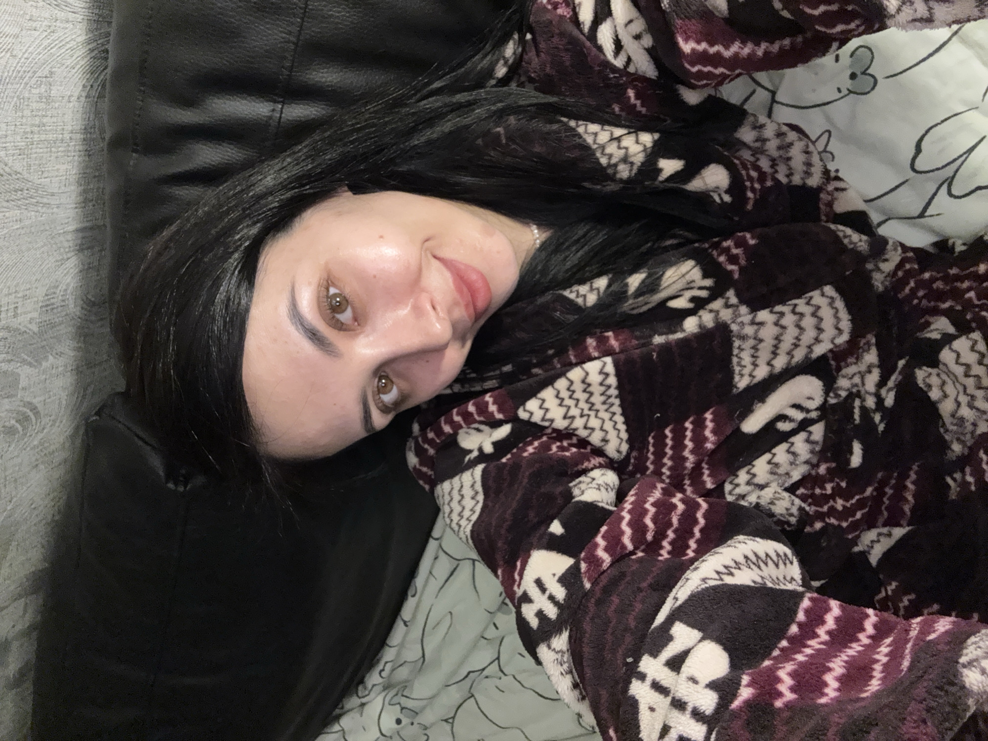 A person is lying on a black leather couch in a room with patterned bedding, wearing a brown and white robe with a zig-zag pattern. The person has black hair and light brown eyes. The location is unspecified within the city of Votkinsk, Russia.