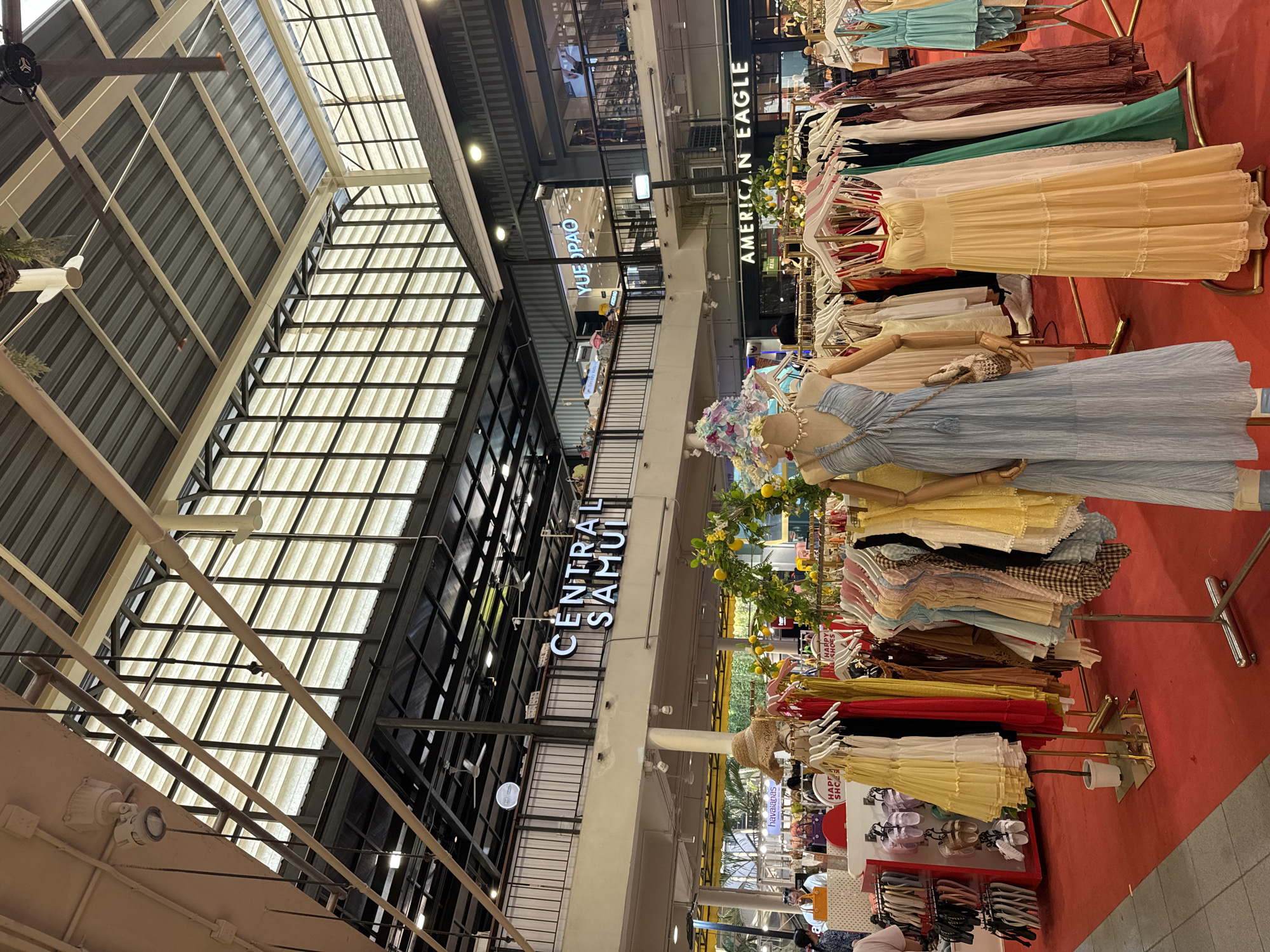 The image captures a well-lit indoor marketplace, likely a shopping mall, in Ko Samui District, Thailand, as suggested by the signage "CENTRAL SAMUI" visible on the upper level. The scene is dominated by racks of clothing, predominantly dresses, displayed on a vibrant red carpeted floor. In the foreground, a mannequin is dressed in a flowing light blue halter dress, adorned with a decorative hat and a small woven bag. Adjacent to it, a pale yellow tiered maxi dress is also displayed. Several other racks of colorful dresses and garments are arranged throughout the space. In the background, more shops and balconies are visible, with prominent signage for "YUE PAO" and "AMERICAN EAGLE". Natural light filters through a large, corrugated roof structure, illuminating the expansive interior. A few shoppers are discernible in the mid-ground, browsing the merchandise. The overall impression is of a lively retail environment.