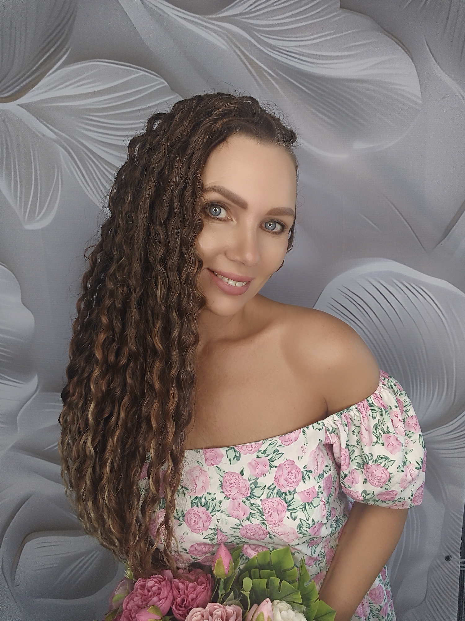 A medium shot photograph shows a woman from the chest up, posed in front of a grey, floral-patterned backdrop. She is looking directly at the camera with a slight smile. Her long, curly brown hair is draped over her left shoulder. She is wearing an off-the-shoulder dress with a floral print of pink roses and green leaves. In the lower right corner of the image, a bouquet of pink and white flowers is visible.