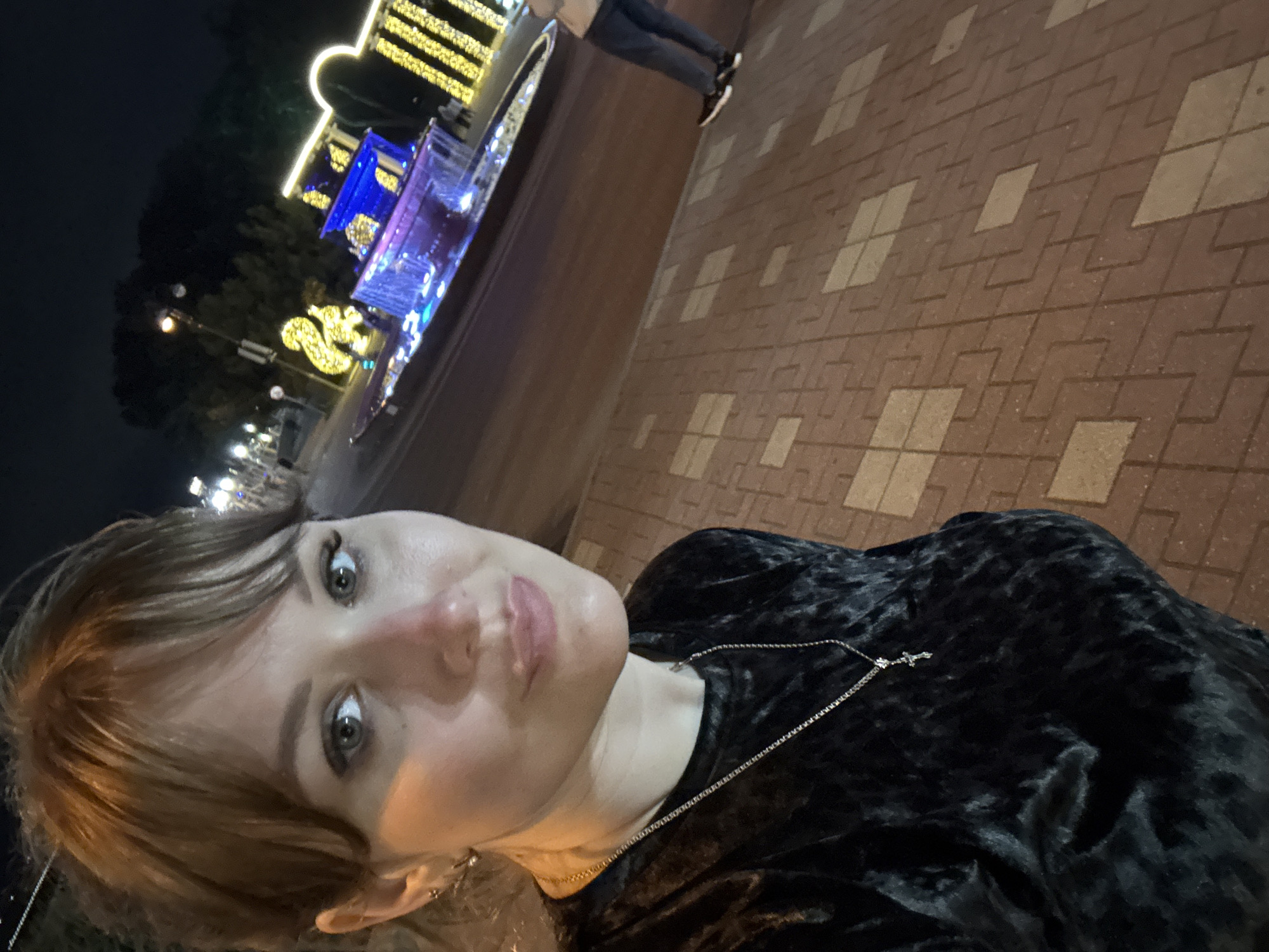 A woman is taking a selfie at night in Gelendzhik, Russia. She is wearing a black velvet top with a silver necklace. In the background, there is a walkway paved with patterned brown bricks. Beyond the walkway, there's a fountain adorned with illuminated decorations, including blue and yellow lights, and some yellow decorative figures. Another person is standing in the background near the fountain.