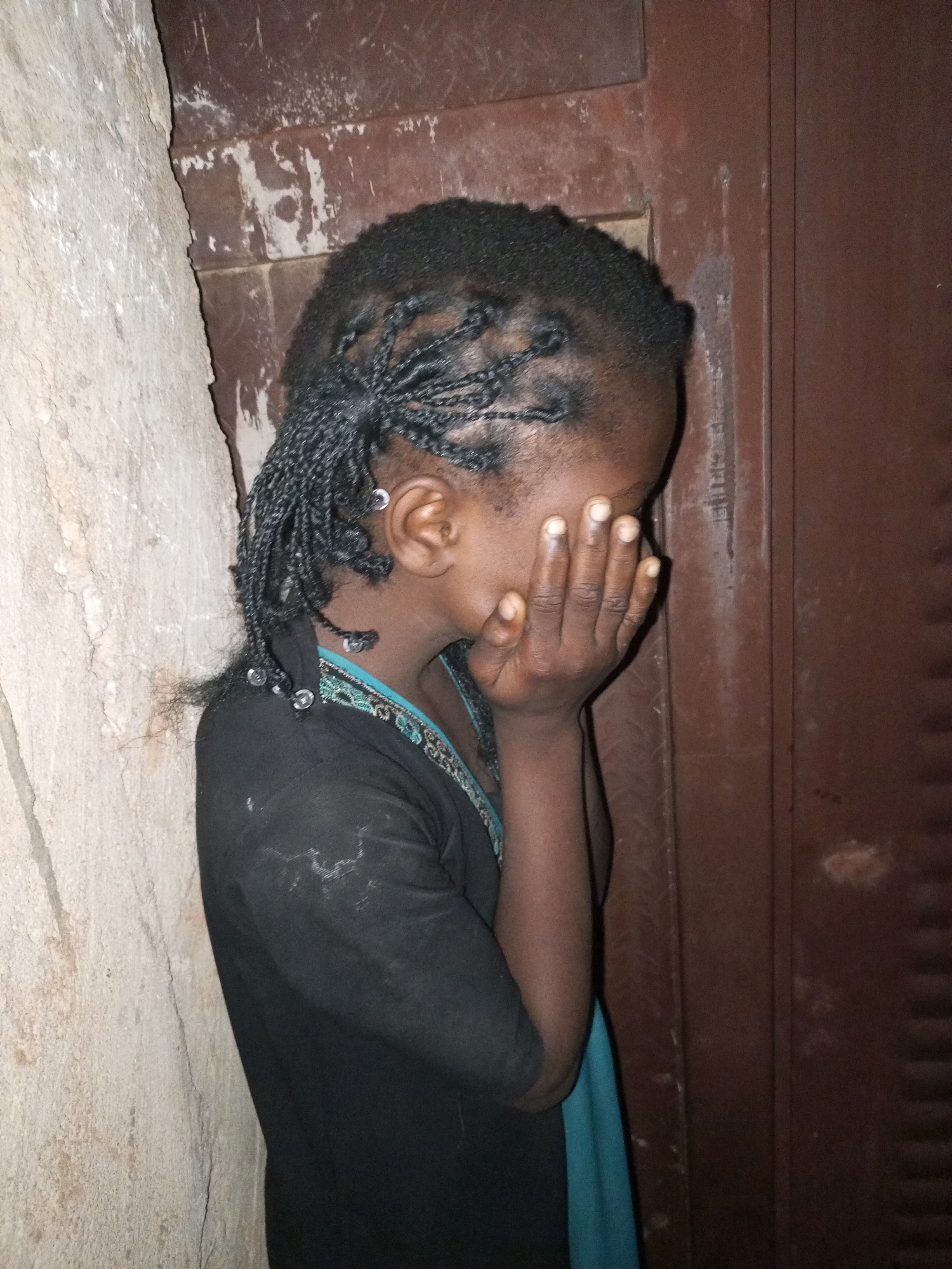 A young Black girl, seen from the side, stands with her right hand covering her face. Her hair is styled in numerous thin braids, some of which are adorned with small, clear beads. She is wearing a black shirt with a teal neckline, and a light blue garment is visible underneath. She is positioned between a rough, light-colored stone wall on the left and a dark brown, textured surface, possibly a door, on the right. The environment appears to be indoors or in a sheltered outdoor area. The image captures a moment of distress or privacy for the girl.