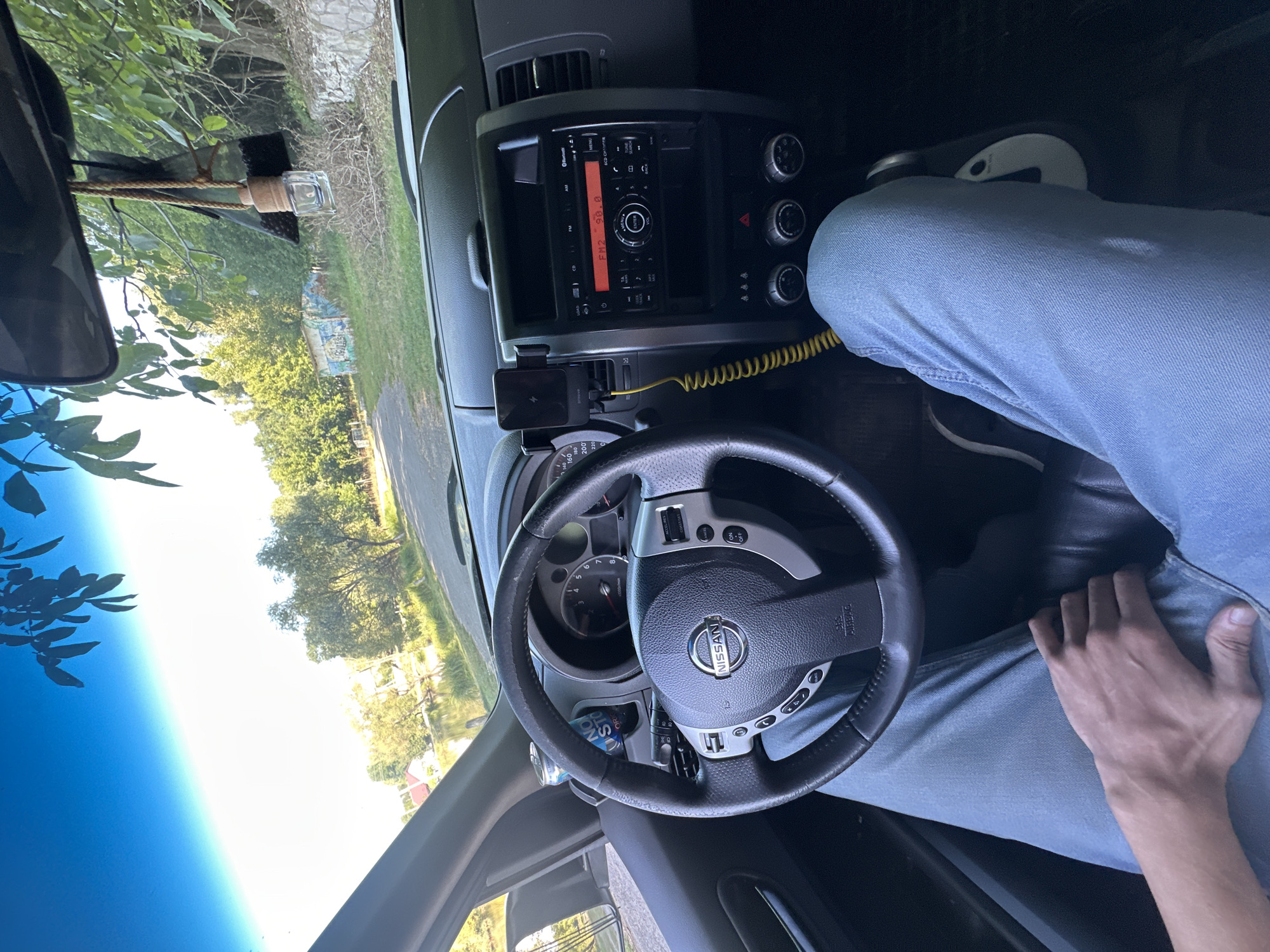 The image shows the interior of a Nissan X-Trail car, looking forward through the windshield. The steering wheel, dashboard, and center console are visible. A person's legs, clad in blue jeans, are in the driver's seat, with their left hand resting on their thigh. A phone is mounted on the dashboard, and a yellow cord is connected to it. The car appears to be parked on a road next to a body of water, with trees and a building in the background. The environment suggests a rural or semi-rural setting. While the general location of Makiv, Ukraine is provided, there are no specific landmarks or identifying features within the image that confirm this location.