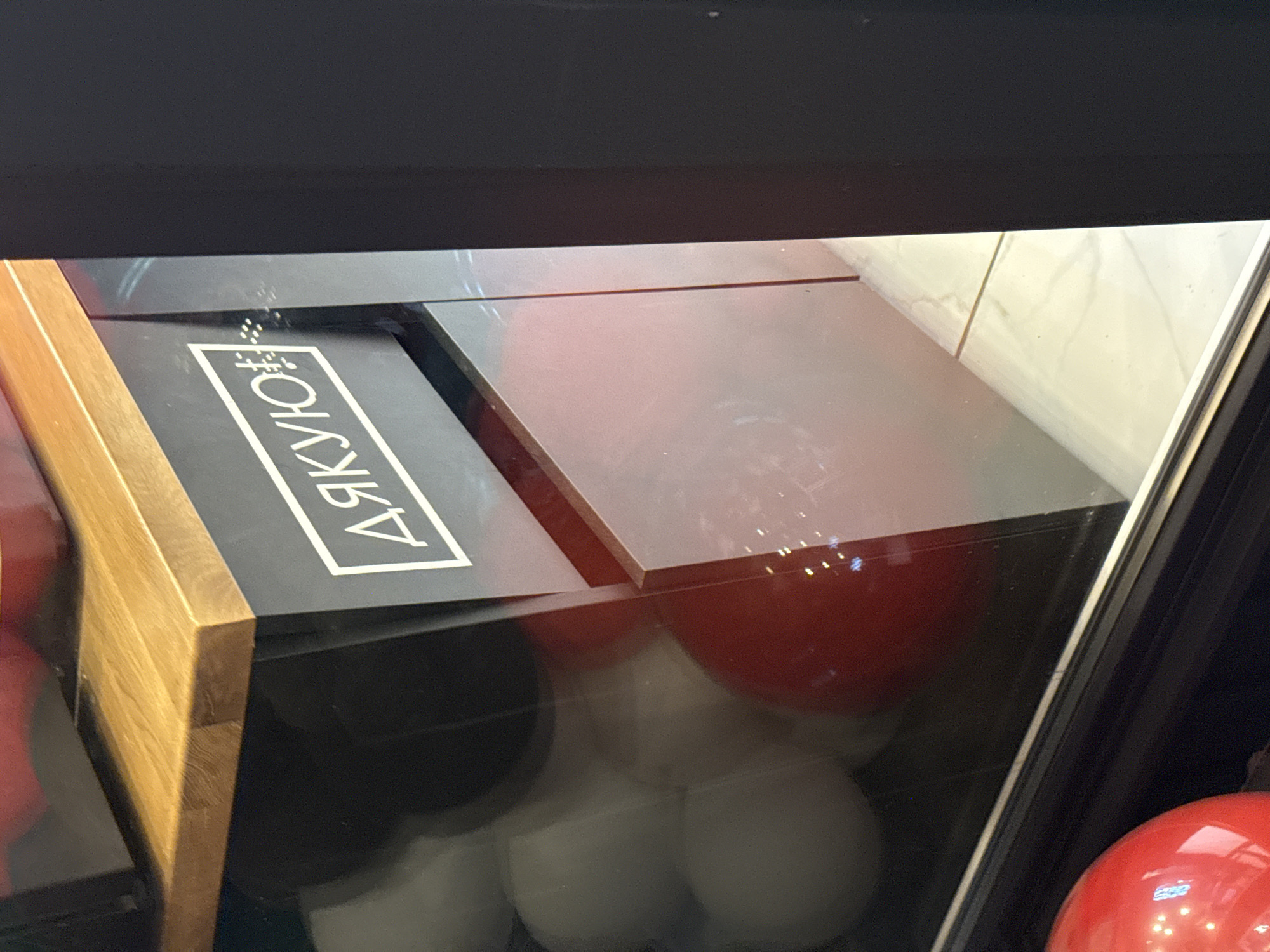 The image shows a close-up view of a black display case with a wooden shelf above it. On the front of the display case, a white rectangular outline contains the Russian word "ДЯКУЮ" (thank you) in white Cyrillic lettering, along with a small symbol that appears to be a coffee cup. The display case has a glass front, through which several round, white objects, possibly balloons or decorative spheres, are visible, as well as a large red sphere in the lower left corner. The background appears to be a tiled floor. The image is taken from a slightly elevated angle, looking down into the display case.