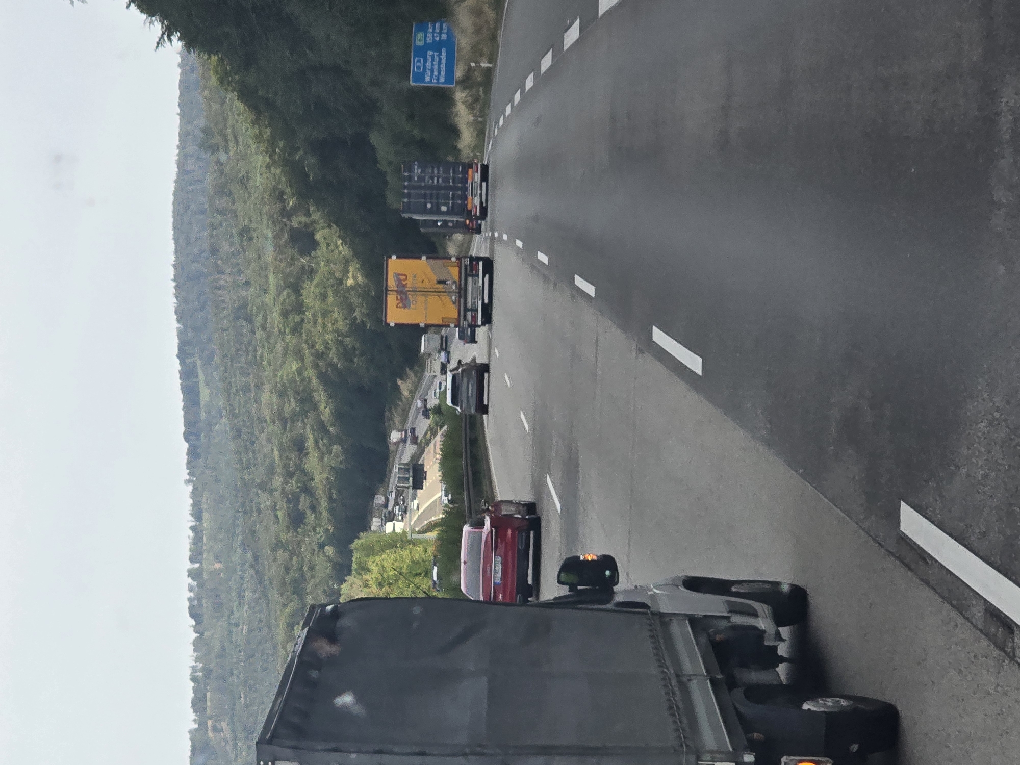 The image depicts a highway scene on a cloudy day, likely in Germany. In the foreground, the rear of a dark-colored truck is visible, obscuring part of the view. Ahead, a red Audi sedan is in the left lane, followed by other vehicles. The right lanes show several trucks, including a yellow one with "DECKO LOGISTIK" branding and a dark container truck. A blue highway sign in the distance indicates distances to Würzburg (158 km), Frankfurt (43 km), and Wiesbaden (18 km). The highway is flanked by dense, green forested hills. The overall impression is one of moderate highway traffic amidst a natural landscape. There is no discernible information to specifically place the scene within Idstein, Germany, although the sign pointing to Frankfurt and Wiesbaden suggests it could be on a route approaching or departing these areas, which are in proximity to Idstein.