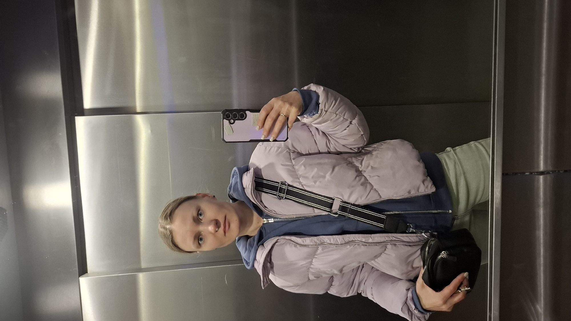 A fair-skinned woman is taking a selfie in what appears to be an elevator with stainless steel walls. She is wearing a lilac puffer jacket over a blue hooded sweatshirt and light green pants. She has a black crossbody bag with a black and white strap. She is holding a lavender phone in her left hand, displaying its rear cameras. The phone case is also lavender. The woman has her hair pulled back, and she is wearing rings on both hands. The lighting is bright, reflecting off the stainless steel. The environment is clean and modern. There is no evidence to support any assumptions about the city being Saint Petersburg, Russia.
