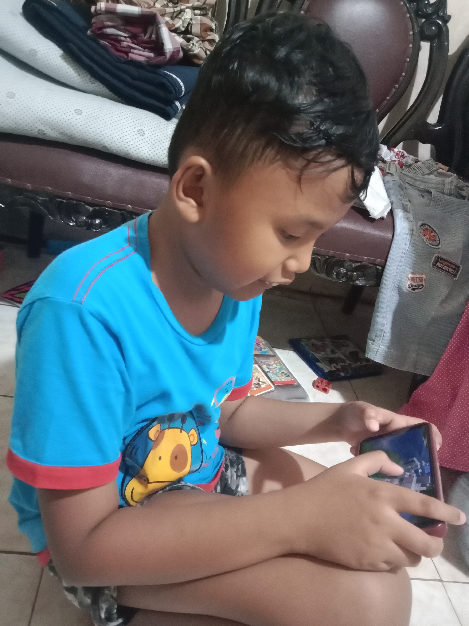 A young boy, appearing to be of East Asian descent, is seated on a tiled floor, engrossed in playing a video game on a smartphone. He is wearing a bright blue t-shirt with a cartoon giraffe design on the front and red trim on the sleeves. His hair is dark and styled short.
The background suggests an indoor domestic setting. To the boy's left, there are stacked pillows and folded fabrics, including a patterned textile and dark blue material, placed on a dark brown upholstered surface, possibly a sofa or chair. Behind him and to his right, a portion of a dark brown wooden chair with carved details is visible, along with a piece of light blue fabric adorned with patches, one appearing to be a circular emblem and another rectangular with text that might read "BRITISH DESIGN" and "LEGENDARY MOTORCYCLE". A pink dotted fabric is also visible on the right side.
On the floor in front of the boy, a stack of what appear to be trading cards or game cards is laid out, alongside a blue rectangular object and a small red brick. The lighting is dim, casting soft shadows. The overall impression is a casual moment of a child engaged with technology within a home environment. This scene could be located in Madiun, Indonesia, given the provided location context.