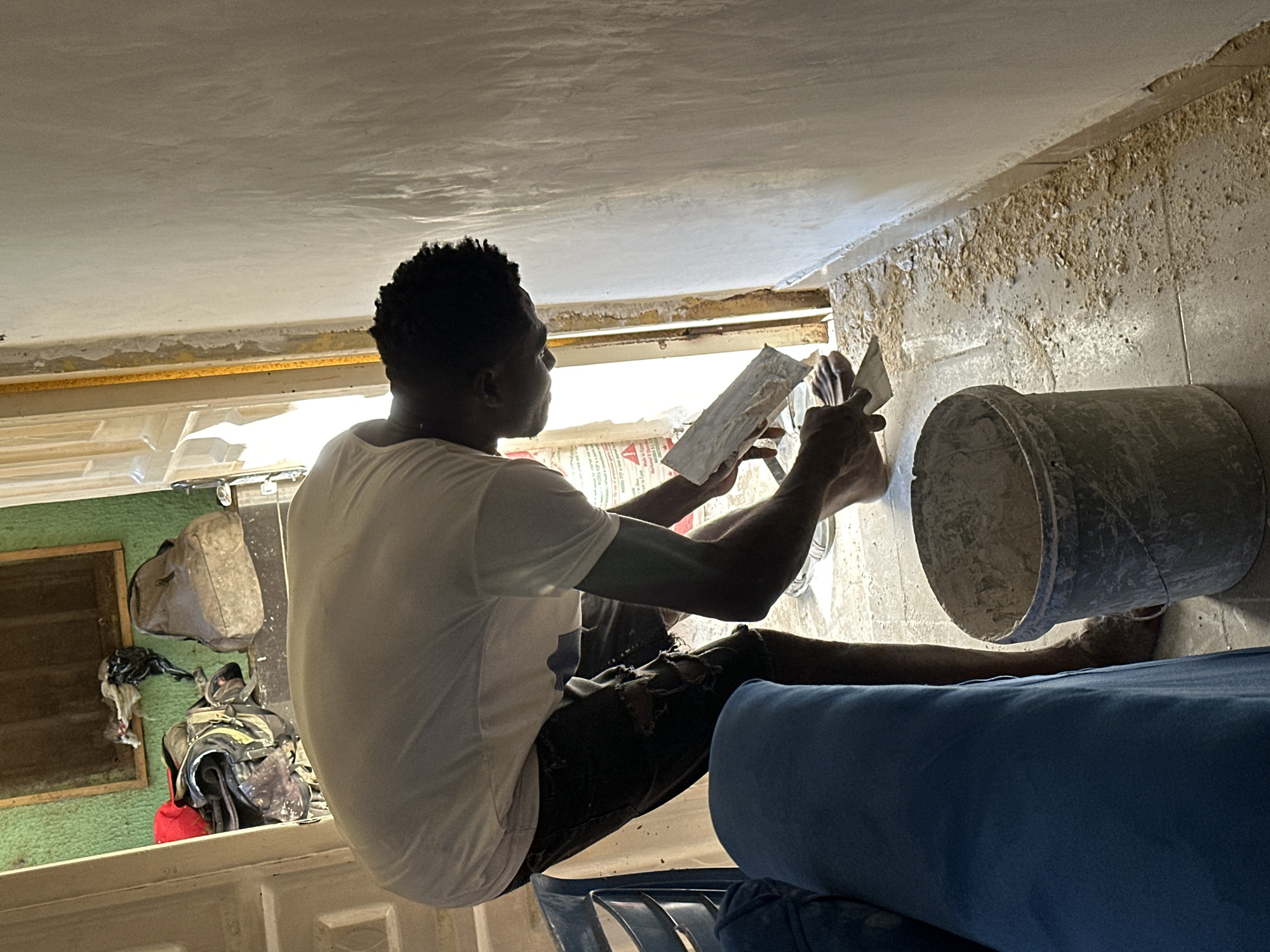 The image captures a person applying plaster to a wall in a room. The person, wearing a white t-shirt and torn dark shorts, is bent over, holding a trowel and applying plaster to the wall with their right hand. Their left hand is also near the wall, possibly holding another tool or steadying the trowel. A bucket containing a plaster-like substance is positioned on the floor to the person's left. Debris, likely plaster fragments, is scattered on the tiled floor around the working area. In the background, an open doorway reveals a hallway with a green wall and several bags and clothing items placed near a window. A blue sofa is partially visible in the foreground on the left. The overall setting appears to be an interior space undergoing renovation or construction. The location is identified as Narayi, Nigeria.