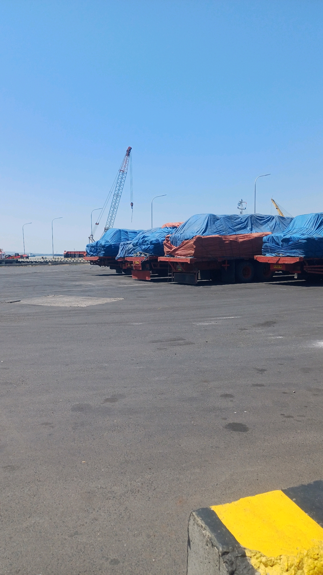 In the city of Lumpur, Indonesia, a wide paved area, likely a port or industrial yard, is captured under a clear blue sky. The ground is a dark asphalt surface with some scuff marks and a concrete barrier painted with yellow and black stripes in the foreground. In the middle ground, a line of several large trucks is parked, their cargo covered with bright blue tarpaulins and some orange sheeting underneath. These trucks appear to be semi-trailers. In the background, a prominent red and white lattice tower crane stands tall against the sky, its hook visible. To the right of the crane, another yellow crane is partially visible. Further in the distance, more industrial structures and what appears to be a body of water with some vessels can be seen. The scene suggests a logistics or shipping hub. There are no people visible in the image.