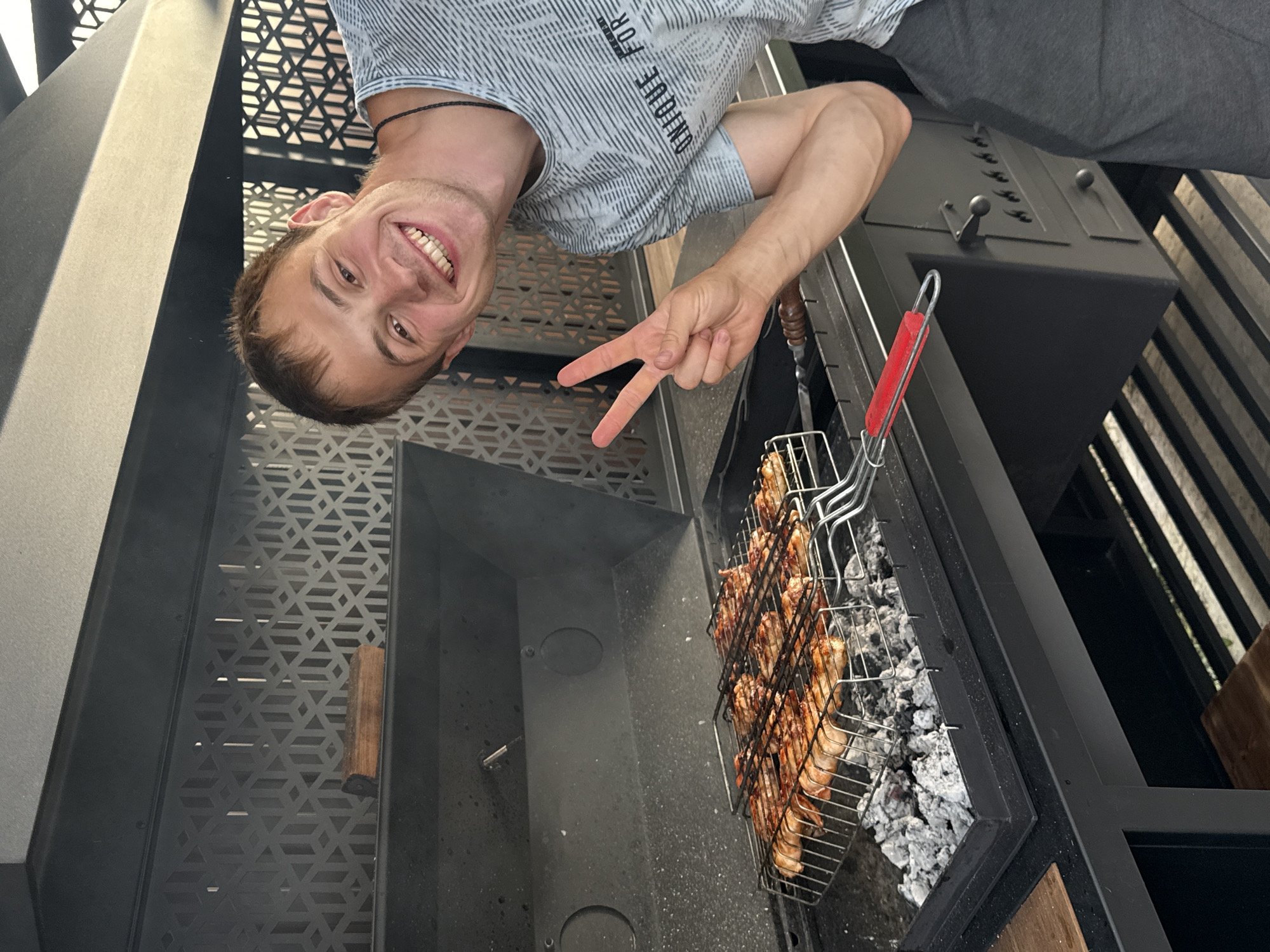 The image shows a young man in Starokorsunskaya, Russia, smiling and making a peace sign while grilling chicken. He is wearing a grey t-shirt with a striped pattern and text that reads "UNIQUE FOR". The chicken is in a grilling basket over hot coals, and smoke is visible. The man is positioned in the foreground, with the grill and a portion of a black outdoor kitchen unit behind him. The overall scene suggests a casual, outdoor cooking activity. The lighting indicates it's daytime.