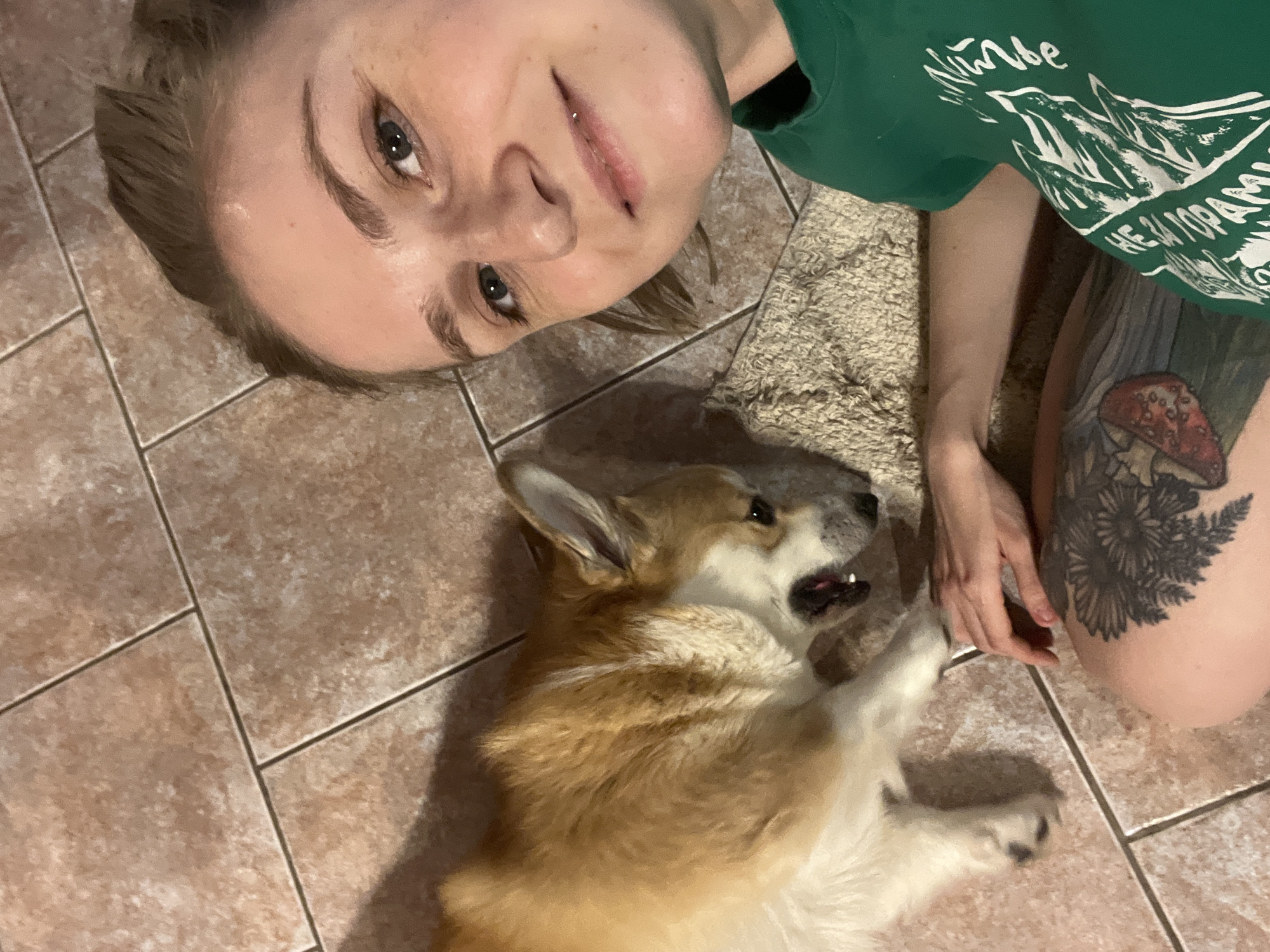 An overhead shot captures a person and a Corgi dog relaxing on a tiled floor, possibly indoors in Novosibirsk, Russia. The person, a light-skinned female, lies on her back with her head towards the top of the frame, looking directly at the camera with a relaxed expression and a slight smile. She has light brown hair, dark eyes, and a lip piercing. She wears a dark green t-shirt featuring white Cyrillic text and graphics, including "НЕ ЗАГОРАЙ" (Don't sunbathe) and what appears to be a mountain or forest design. A large, intricate black tattoo depicting a red mushroom (likely a fly agaric) and fern-like plants is prominent on her upper left thigh.
Beside her, a brown, white, and black Corgi dog also lies on its back, appearing content or playful with its mouth slightly open. Its gaze is directed towards the person's hand, which rests near the dog's paw. A textured, light brown rug or mat is partially visible between the person and the dog. The floor is covered in light brown or beige tiles with dark grout lines. The image conveys a moment of affectionate interaction and relaxation between the owner and their pet.