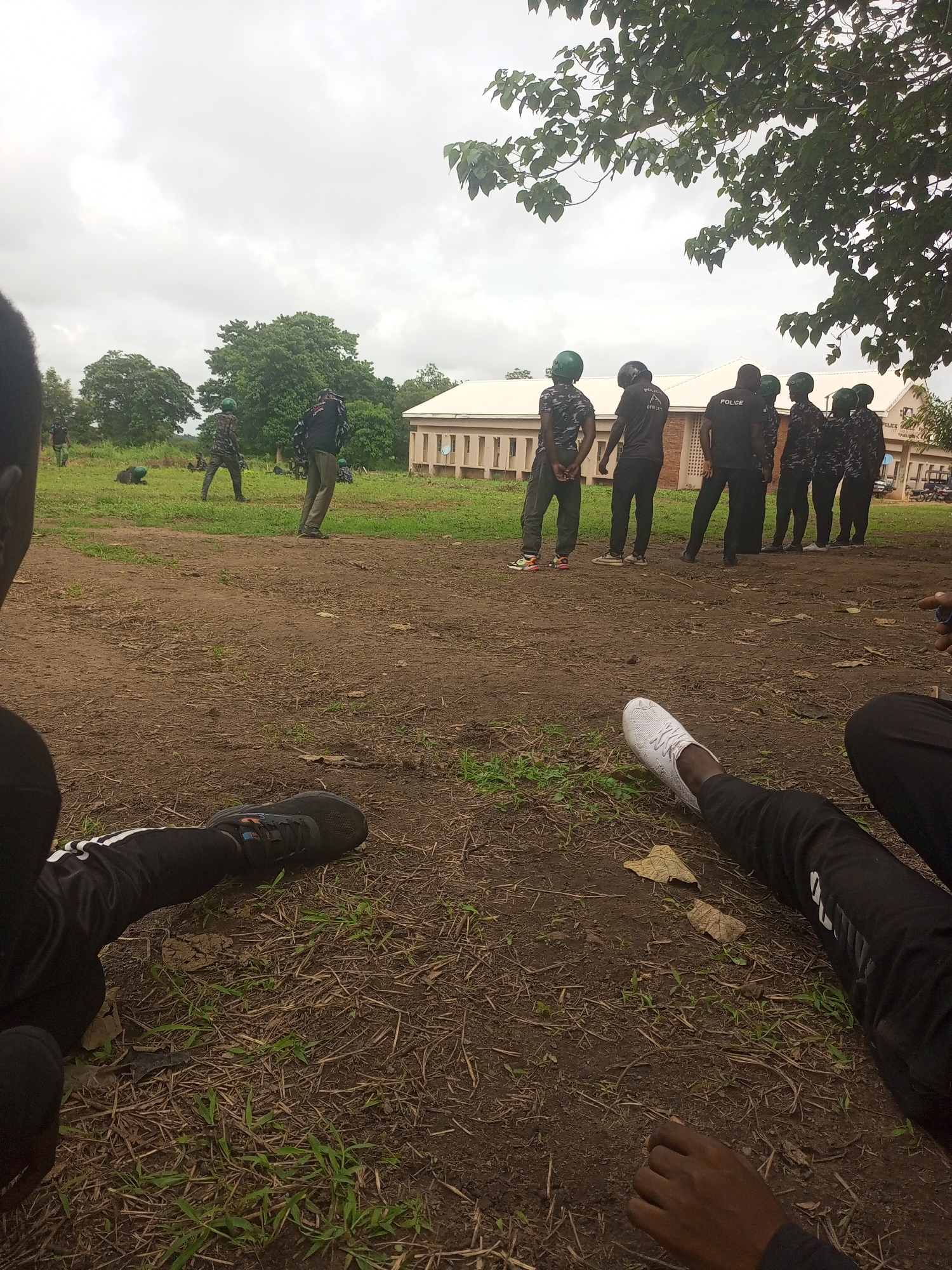 The image depicts an outdoor scene, likely in Takum, Nigeria, with a group of individuals, predominantly young men, in what appears to be a training or drill exercise. In the foreground, two individuals are partially visible, with one wearing black athletic pants with white stripes and a black shoe, and another with a white athletic shoe and black pants. In the middle ground, a larger group of men are standing in formation, all wearing dark camouflage-patterned uniforms with green helmets. Some of these individuals have "POLICE" written on their shirts. Their stances suggest they are engaged in a formal activity. Further in the background, a long, light-colored building with a tiled roof is visible. The surrounding environment is grassy and somewhat muddy, with trees in the distance and a cloudy sky overhead. There are also some individuals in camouflage uniforms further back in the field, with one person appearing to be in a prone position. The overall impression is of a disciplined gathering, possibly a police or security force training session.