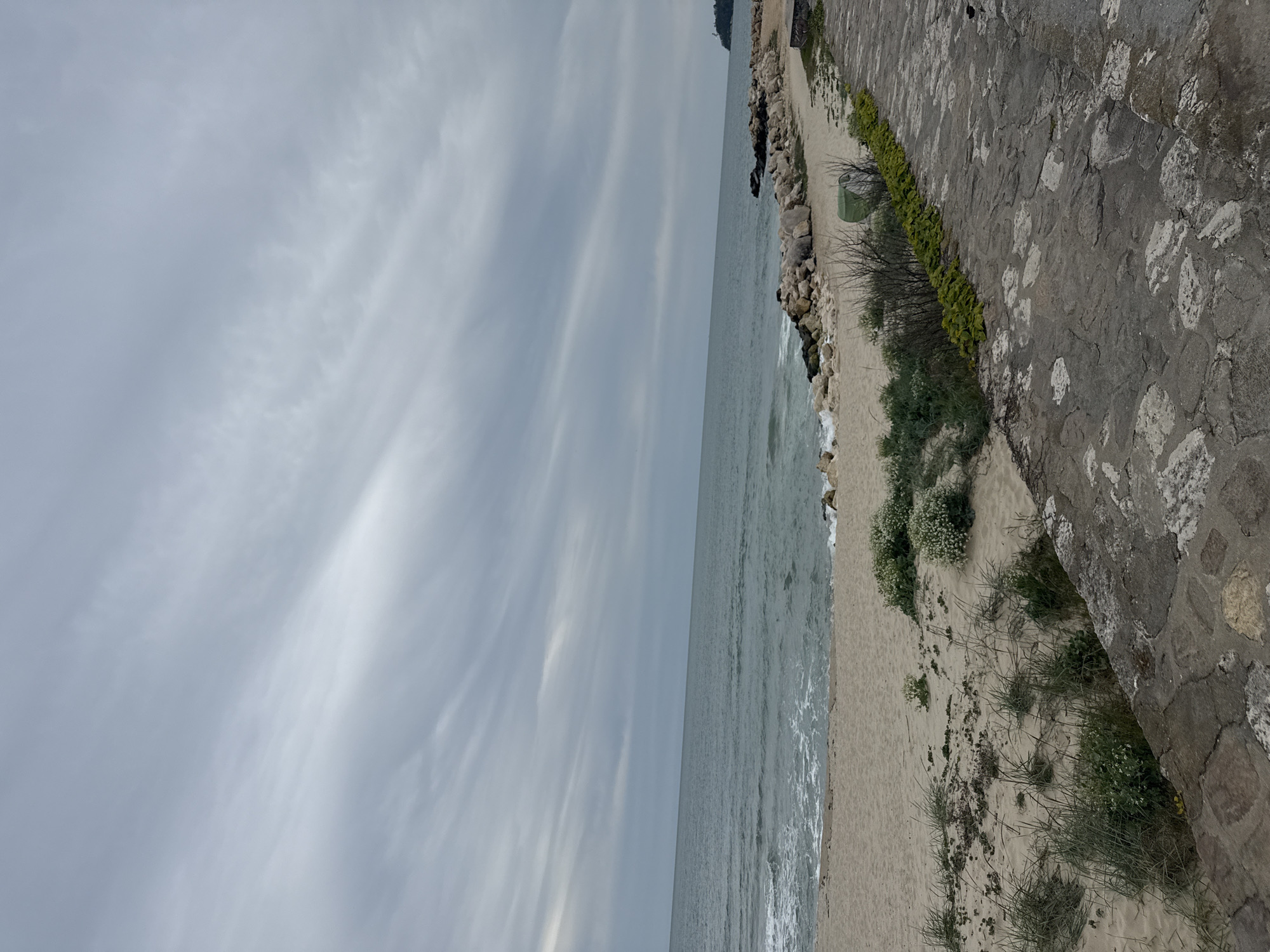 The image depicts a coastal scene in Varna, Bulgaria. A stone wall borders a sandy beach. The sea is visible along the coastline. The sky is overcast with striated clouds. No people are visible.