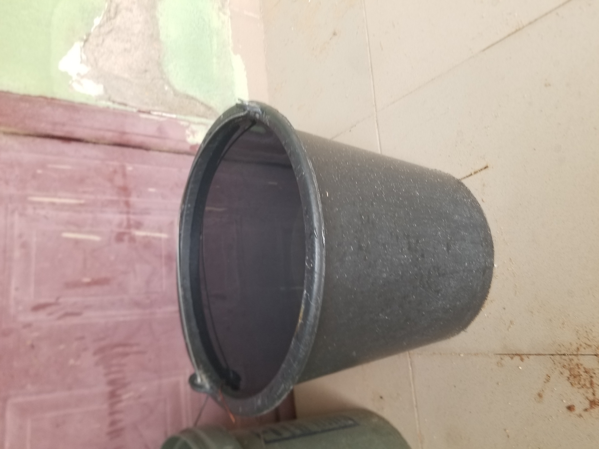 A black plastic bucket containing water is positioned on a tiled floor. The bucket is centrally located in the frame and is a dark, speckled color. The water inside reflects the surroundings. To the left of the bucket, partially visible, is another green plastic container. Behind the bucket, a wall with a reddish-brown section and a lighter green section is visible. The floor tiles are light-colored and appear to be laid in a grid pattern. There are some scattered brown specks on the floor, possibly dirt or debris, particularly near the base of the bucket and to its left. The scene is indoors or in a sheltered outdoor area, with the wall suggesting a building. The image is shot from a slightly elevated angle, looking down towards the bucket. This scene is located in Jalingo, Nigeria.