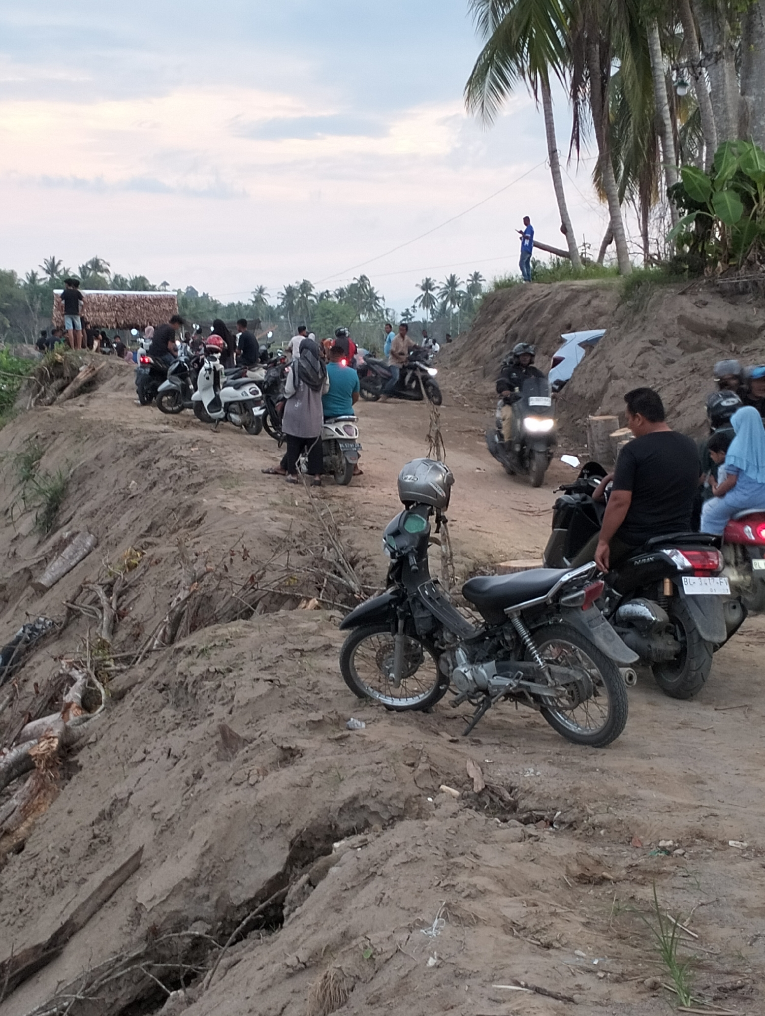 A crowded scene unfolds at what appears to be a coastal location, possibly a tourist destination in Kapa, Indonesia. Numerous individuals and motorcycles are congregated on a dirt road or embankment. The ground is eroded, exposing underlying soil and tree roots.
People: Many people are present. They are diverse in age and appearance, mostly dressed casually. Some are on motorcycles, others are standing and interacting. One man is positioned on an elevated section near palm trees.
Subjects: Motorcycles of varying types and conditions are a central element, indicating a common mode of transportation and congregation.
Objects: Besides motorcycles, other identifiable objects include palm trees and a structure with a thatched roof.
Actions: People are congregating, possibly observing something out of frame, riding motorcycles, or simply enjoying the location.
Interactions: The scene suggests a social gathering. Interactions include people standing together, some on motorcycles interacting with others, and a general sense of collective activity.
Environment: The setting is coastal with palm trees dominating the backdrop. The terrain is uneven with apparent erosion of the ground. The time of day appears to be late afternoon or early evening, indicated by the lighting.