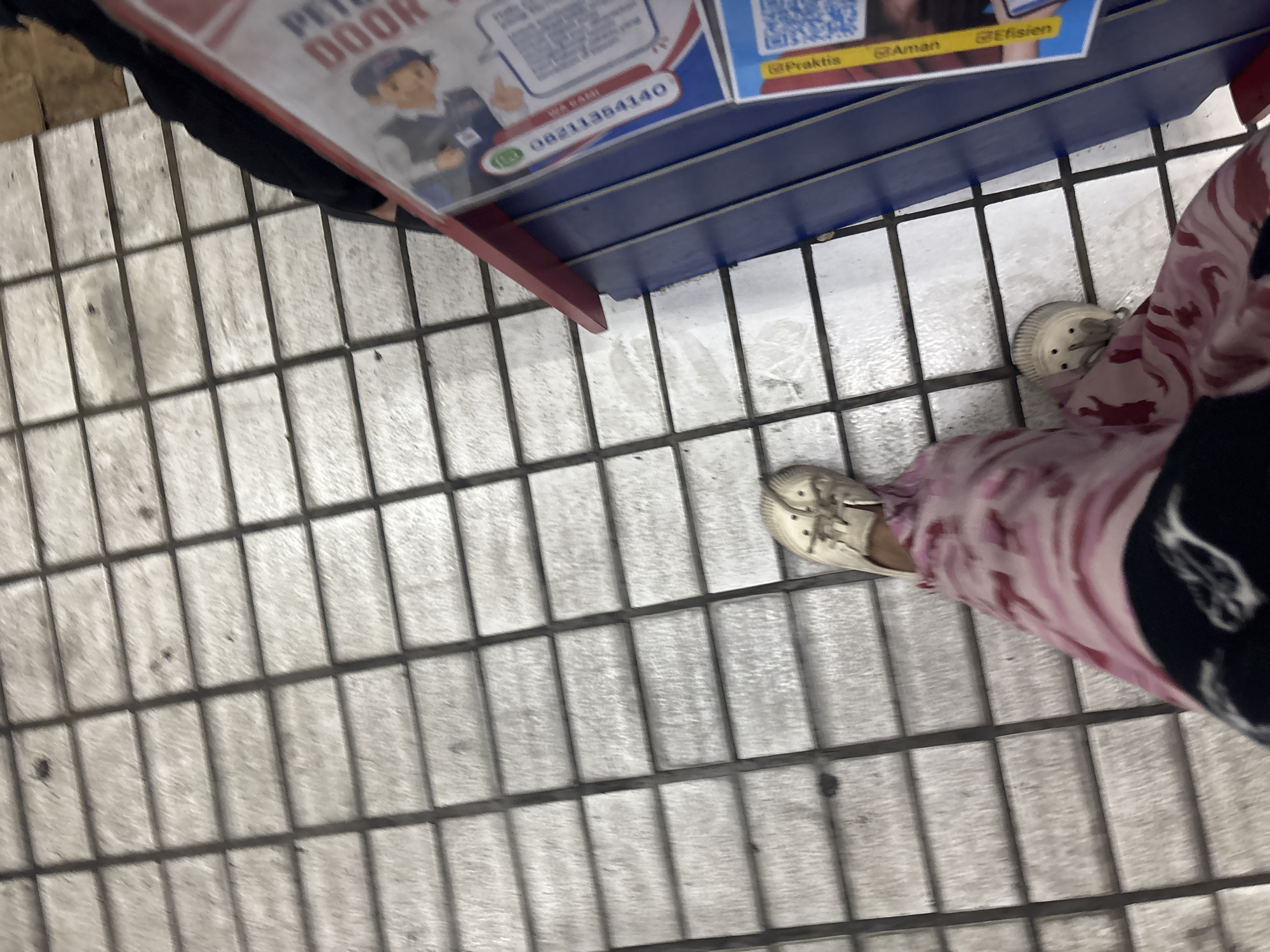 The image shows a person's legs and feet from a top-down perspective, standing on a tiled surface. The person is wearing a pink and white patterned skirt or dress and off-white clogs. To the right of the person, there is a blue and red display with some text and images on it. The text on the display appears to be in Indonesian. The tiled surface is made of rectangular tiles, arranged in a grid pattern. Some areas of the tiles appear slightly wet. The overall scene suggests an outdoor urban environment. There are no visible landmarks to pinpoint a specific location within North Jakarta, Indonesia, but the presence of a display with Indonesian text suggests it is indeed in Indonesia.