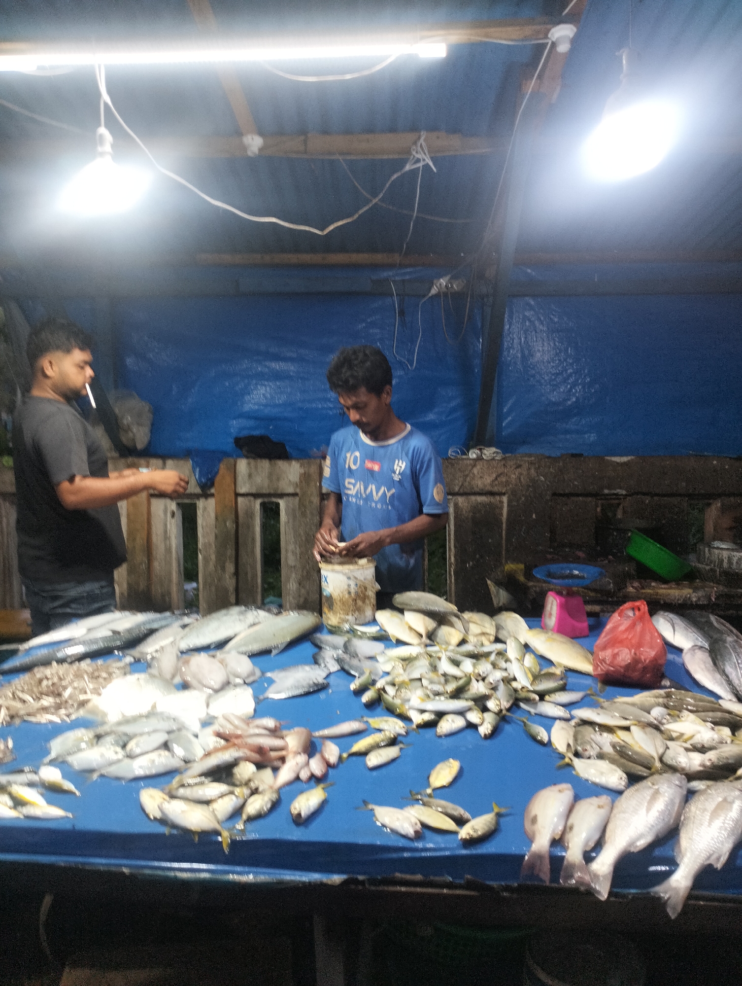 The image shows a fish market stall, likely in Lhokseumawe, Indonesia. Two men are present. The man on the left is holding a cigarette. The man on the right is wearing a blue shirt and working with a container, possibly cleaning fish. A table in front is covered with a variety of fish, including small yellow fish and larger silver fish. A red plastic bag and a pink scale are on the table. The stall has a blue tarp as a backdrop, and two bare light bulbs illuminate the scene. The stall appears to be partially open to the outdoors.