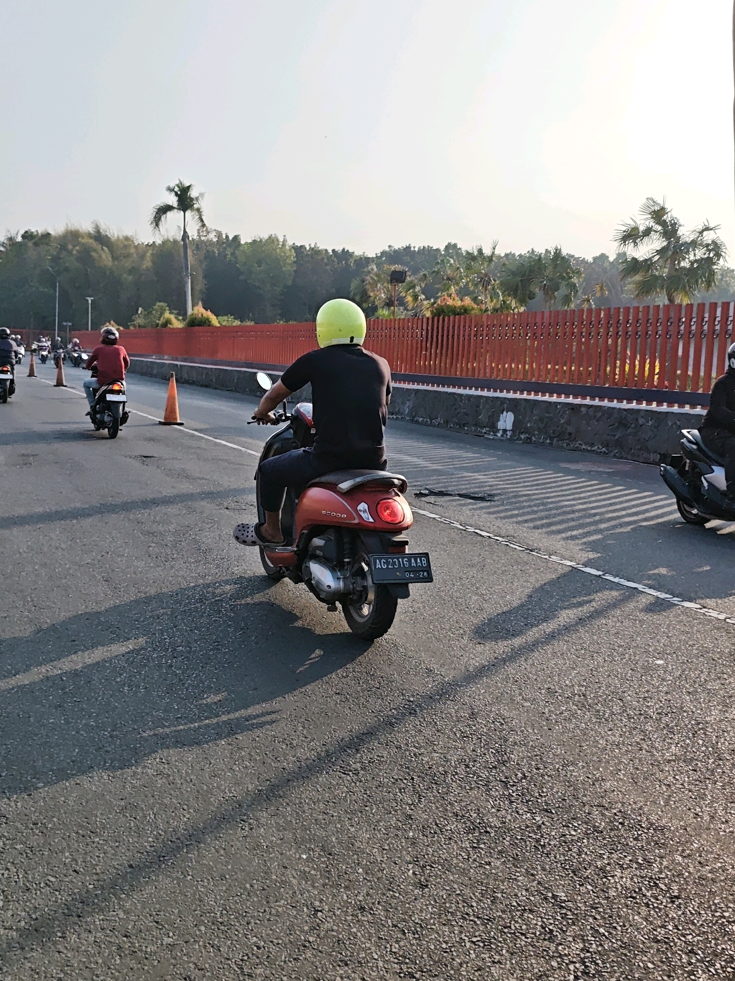 A group of motorcyclists ride on a road in Kediri City, Indonesia. The road has a white line marking and is bordered by a red fence and trees. The sky is bright and clear. A man in a yellow helmet rides a red scooter in the foreground, his license plate reading "AG 2316 AAB". Other motorcyclists are visible in the background, some wearing red and black clothing. Orange traffic cones are placed on the side of the road. The scene appears to be during the daytime, with shadows cast on the asphalt from the motorcycles and the fence.