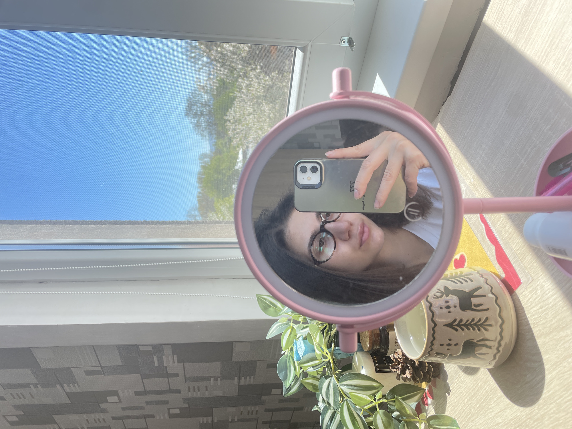 The image shows a selfie taken in a mirror. The reflection includes a person with dark hair, wearing glasses, holding a smartphone. The phone has a gray case and a dual-lens camera. The person is wearing a white shirt. The mirror is pink and round, standing on a table.
On the table, near the mirror, are a ceramic cup with deer and tree patterns, a plant with green and white striped leaves, a pinecone, and various bottles and containers. A yellow card with a red heart on it is also visible.
Behind the mirror is a window with a view of a clear blue sky and trees with white blossoms. The window has a roller blind. The wall below the window has a gray geometric pattern. Based on the style of the window, and the architectural style of nearby buildings, it is likely that this picture was taken somewhere in the city of Sofia, Bulgaria.