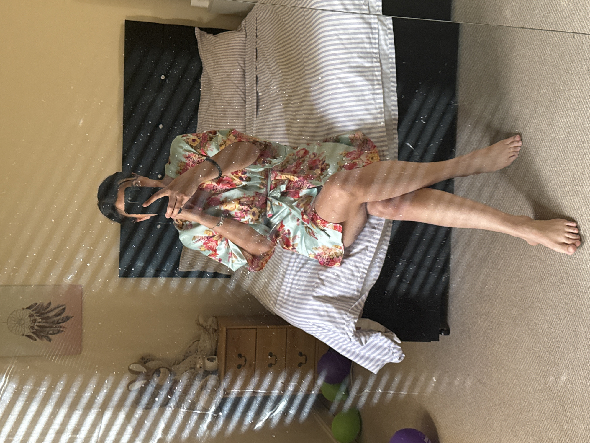 A person is sitting on a bed in a bedroom, reflected in a mirror. The person is wearing a floral robe and holding a smartphone. The bedroom has a bed with striped bedding, a bedside table with a stuffed animal and a dreamcatcher on the wall, and balloons on the floor. Sunlight streams through blinds, casting shadows on the wall and floor. The scene takes place in a bedroom in Great Yarmouth, United Kingdom.