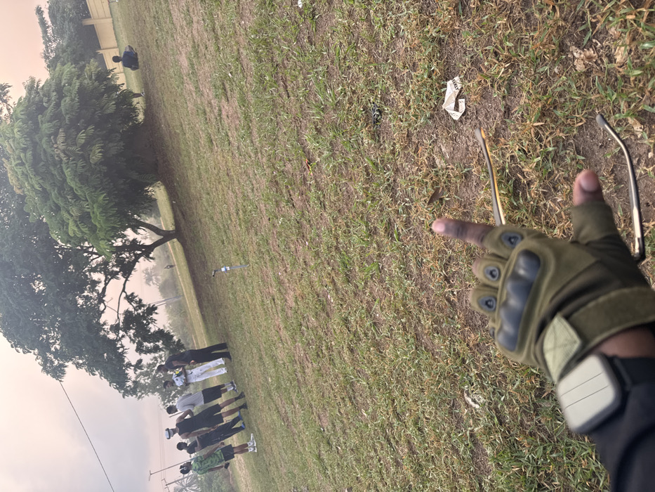 The image captures an outdoor, grassy area in Obinze, Nigeria, under a hazy sky with a pale orange tint. In the immediate foreground, a person's right hand, clad in an olive-green fingerless tactical glove featuring black knuckle protection and a light-colored smartwatch, extends with the index finger pointed upward. A pair of sunglasses is partially visible, draped over or near the hand. The ground is covered with a mix of green and dry brown grass, interspersed with patches of bare earth and scattered debris, including a crumpled white wrapper. A light blue, slender pipe or post protrudes from the ground in the mid-left. In the left background, a group of approximately seven males stands on the grass, casually dressed in various shirts and pants/shorts, appearing to be in conversation or observing their surroundings. In the right background, a large, dense green tree stands prominently. To its right, an individual is seated on the ground near a light-colored wall or fence, partially obscured by the tree. Distant power lines and additional foliage are visible along the horizon.