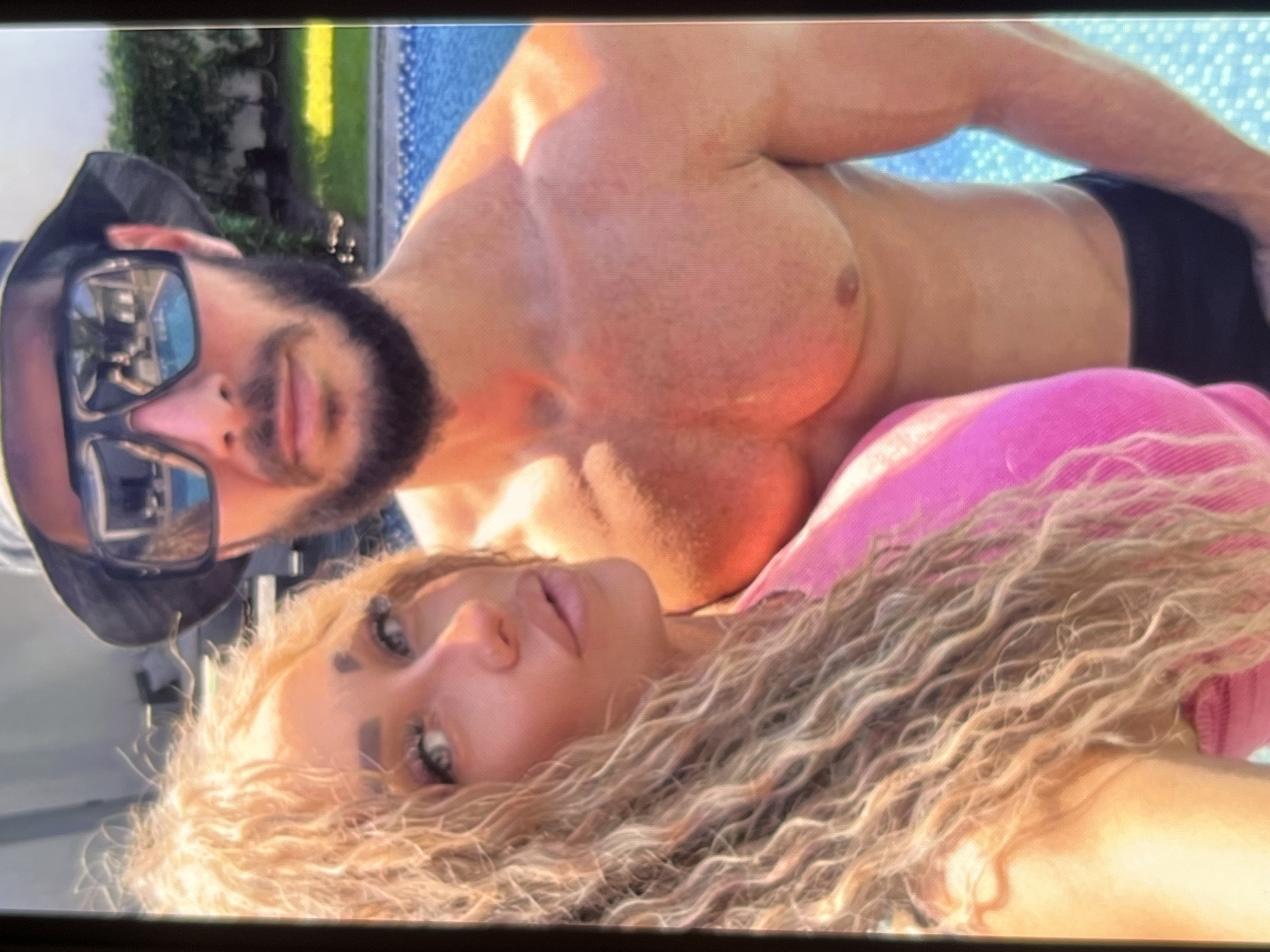 A close-up, medium shot captures a couple posing by a swimming pool. The woman, in the foreground on the left, has long, blonde, curly hair and is wearing a pink top. She is looking towards the camera with her head tilted. Beside her, a man in the background wears a bucket hat and large, dark sunglasses. He has a beard and is shirtless, with his chest and arms visible. The man is looking forward, and his sunglasses reflect the surrounding environment, including a swimming pool and some greenery. The background shows a blue tiled swimming pool and a lush green hedge. The lighting suggests it is a sunny day.