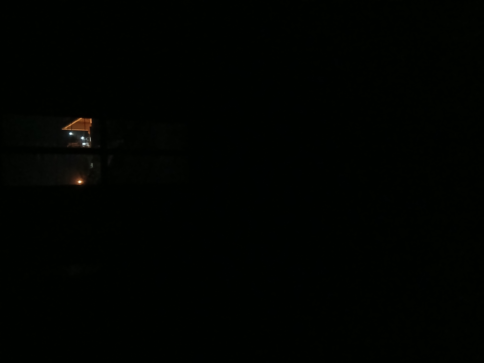 The image is a night-time photograph taken from inside a dark room, looking out a window. The view outside is dimly illuminated by artificial lights. In the distance, a building with a distinctive triangular roof structure is visible, highlighted by lights. Several streetlights or illuminated poles are also present in the scene. The overall impression is one of a rural or semi-urban setting at night, with the lights suggesting some level of development. No people or specific environmental details such as vegetation or terrain are clearly discernible due to the darkness and low light conditions. The specific location in Tenggarang, Indonesia, cannot be determined from the visual information provided.