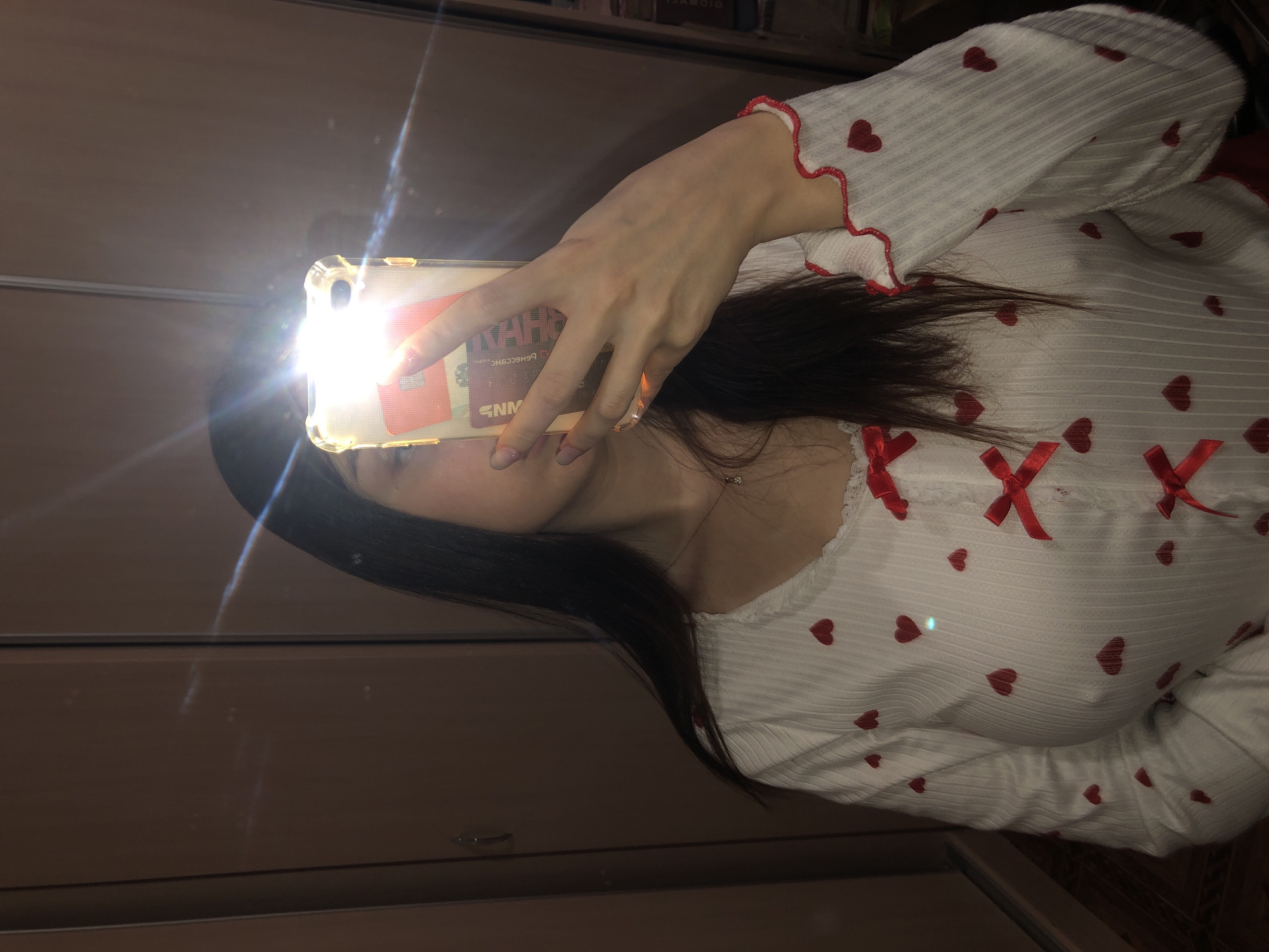 A selfie taken by a person facing left, wearing a white, long-sleeved shirt adorned with red hearts and red ribbons, and holding a phone with a bright flash. The background appears to be a mirrored surface, possibly a closet door. The image does not contain enough information to identify a location in Noviy, Russia.
