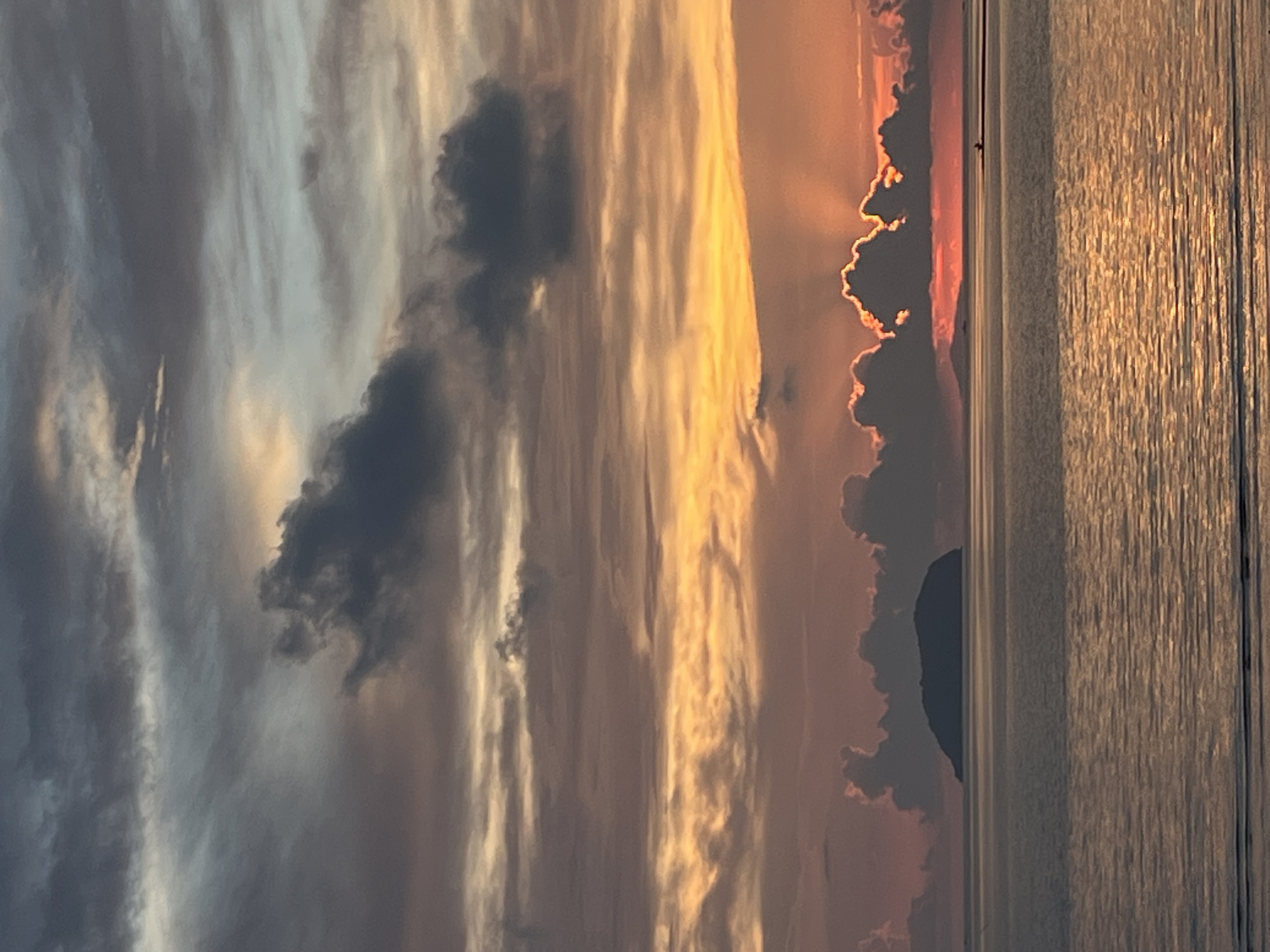 This image captures a serene sunset over the sea near Cuyo, Philippines. A silhouetted island sits on the horizon, its dark shape contrasting with the vibrant sky. The water reflects the warm hues of the sunset, with ripples creating a textured surface. Clouds are scattered across the sky, some dark and dramatic, others illuminated by the setting sun, casting rays of light. A small boat is visible in the distance, adding a sense of scale and tranquility to the vast seascape. The overall scene is peaceful and picturesque, showcasing the natural beauty of the location.