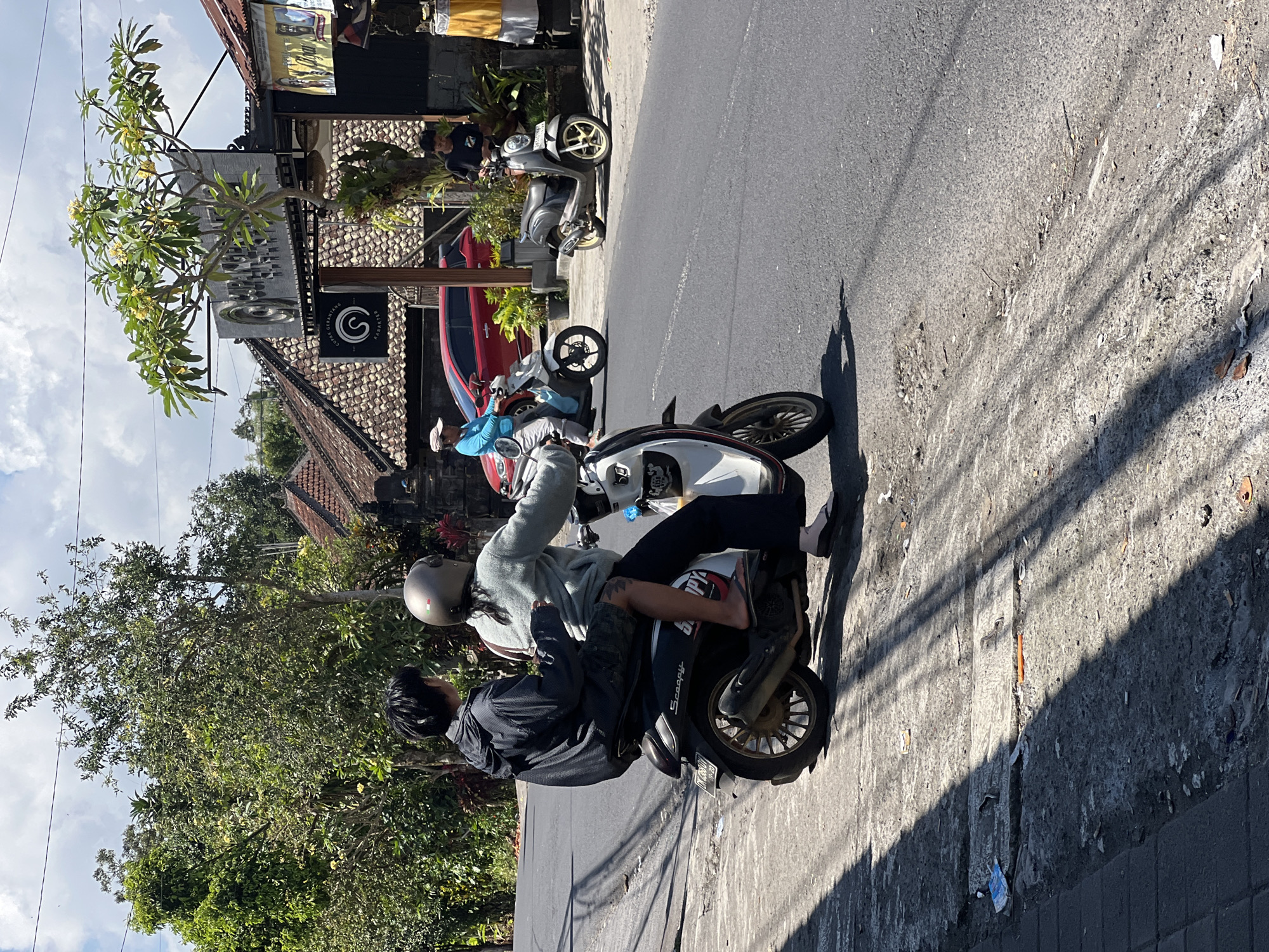 In Payangan, Indonesia, two individuals are riding a scooter on a paved road under a cloudy sky. The person in front is wearing a light blue sweatshirt and a helmet, while the person in the back is wearing a black jacket and shorts. They are positioned on the right side of the road, with a row of trees and buildings lining the background. The environment appears to be a tropical setting with lush vegetation. Several other scooters and a red car are visible in the distance, suggesting a moderately trafficked area. The road surface is asphalt with some visible debris and shadows cast by overhead objects, likely trees or power lines. The scene captures a moment of everyday life in this Indonesian town.