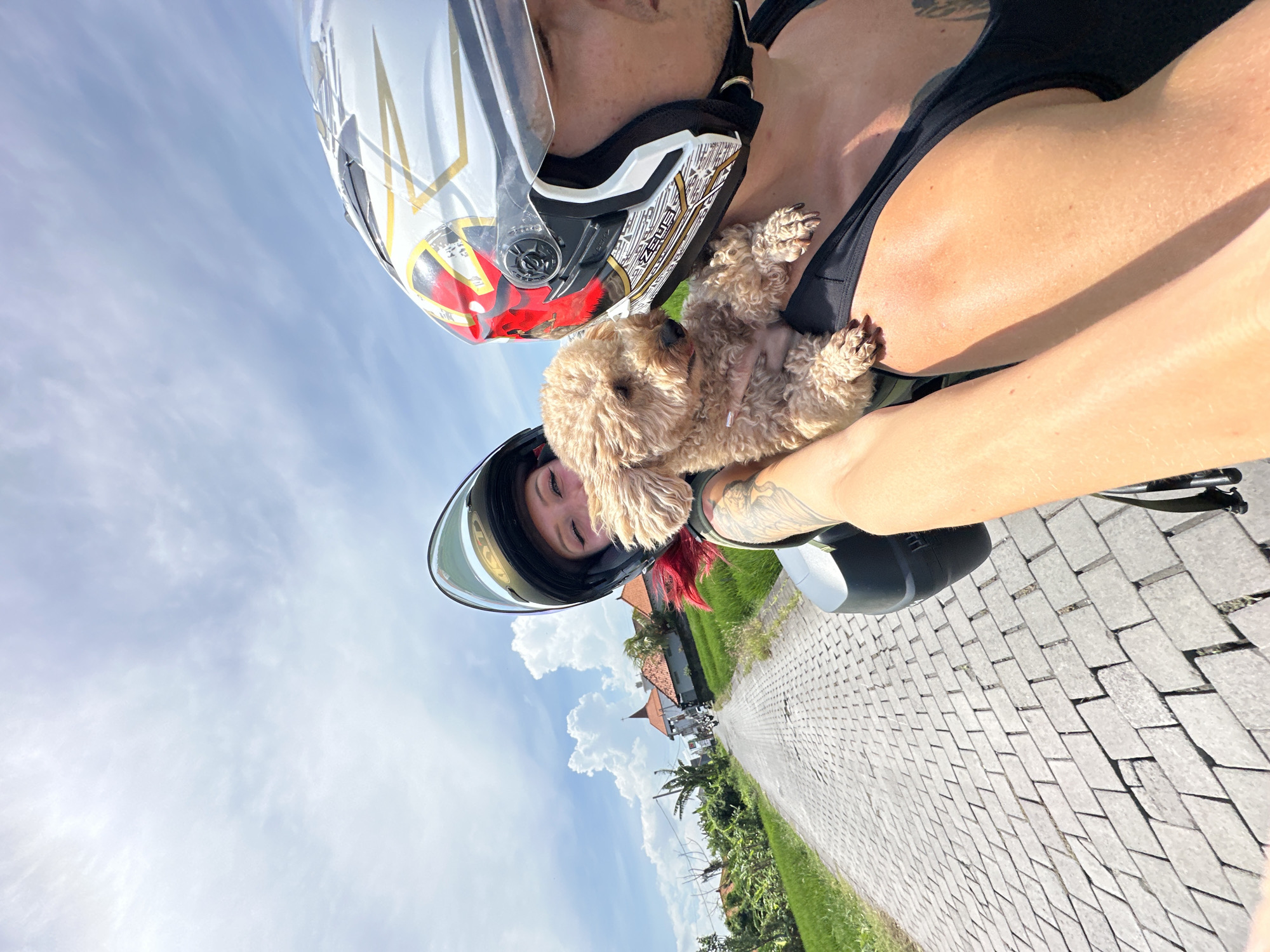 The image depicts a woman wearing a white and gold motorcycle helmet and a black top, holding a small, light-brown poodle-type dog against her chest. The dog is lying on its back. The woman is likely on a motorcycle, as indicated by the handlebars visible on the right. Another person, likely the driver of the motorcycle, is seen below her, also wearing a motorcycle helmet.
The scene appears to be outdoors on a paved road with greenery on either side. Buildings with terracotta roofs can be seen in the background. Based on the location in North Kuta, Indonesia, the image likely captures a ride on a scooter through a rural or semi-rural area. The sky is visible with scattered clouds.