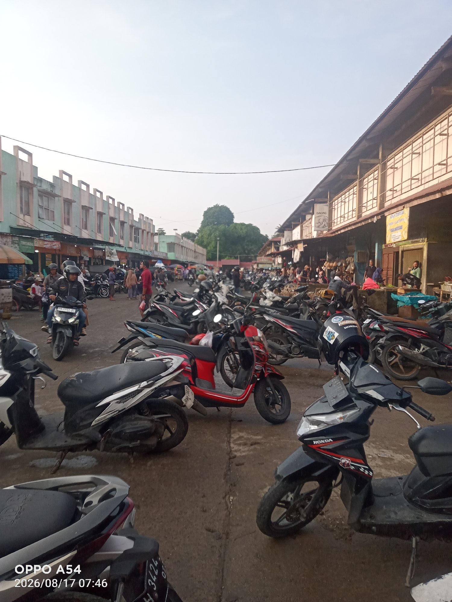 This image depicts a bustling outdoor market scene, likely in the city of Jambi, Indonesia. A prominent feature is a large parking area filled with numerous motorcycles, suggesting the market is a popular destination.
The market itself appears to be an open-air marketplace with stalls and shops lining one side of the street. These structures have a rustic, possibly wooden construction, with corrugated metal roofs. Some of the stalls display goods, including what appear to be food items like meats, judging by a sign that says "DAGING" (meat).
People are visible in the market, some walking and browsing, while others are engaged in transactions or operating their motorcycles. The overall atmosphere suggests a busy commercial day. The road surface is paved but appears somewhat worn and uneven, with patches of wetness or dirt. The sky is bright, indicating daytime, with a hazy or slightly overcast condition. The architecture of the buildings on the opposite side of the market area suggests a more modern, perhaps commercial or residential, development with rows of windows. The presence of a mature tree in the background adds to the urban landscape.