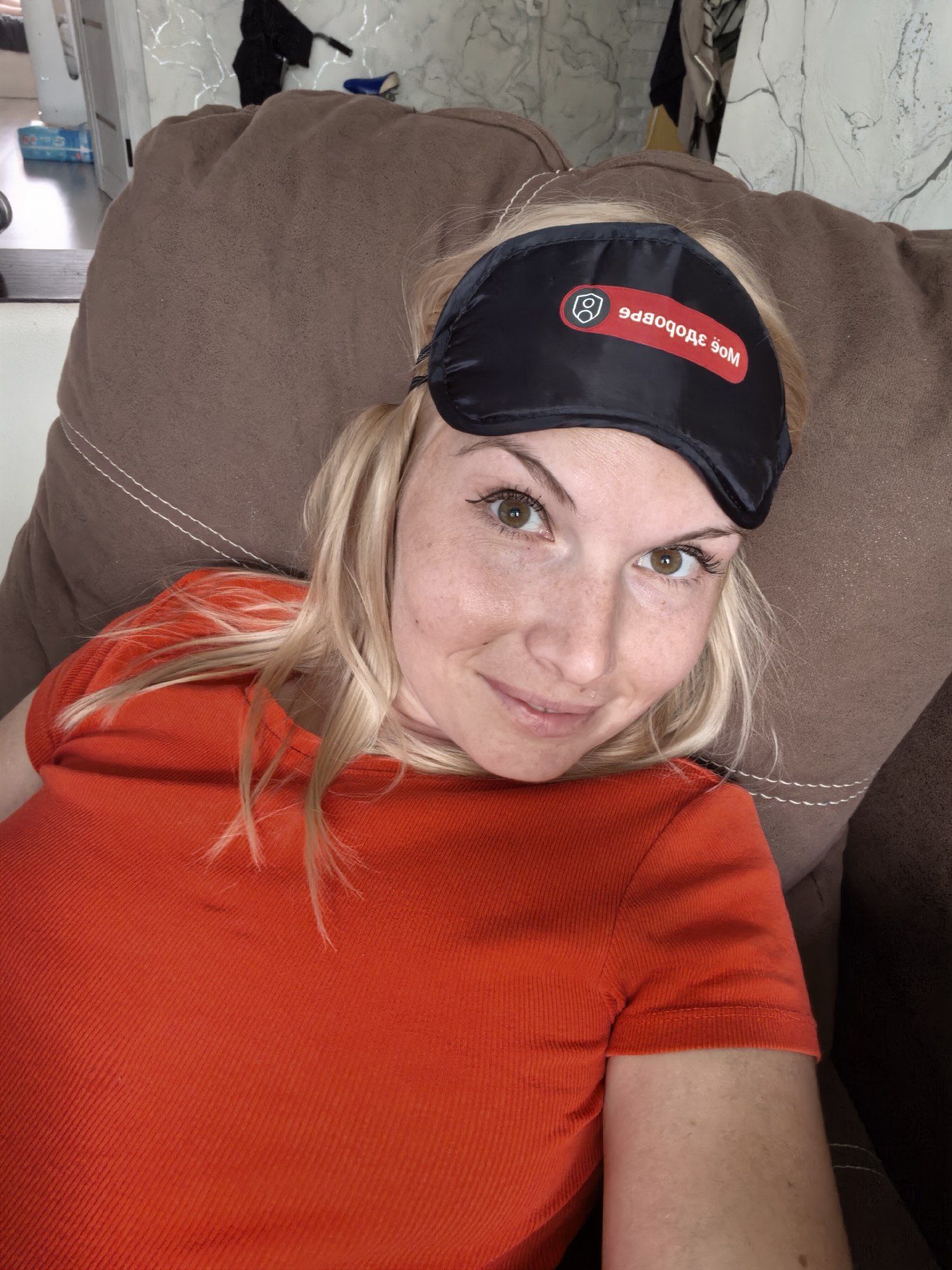 A woman with blonde hair and green eyes is lying down on a brown couch, wearing a black sleep mask with a red logo and an orange t-shirt. The sleep mask has the Cyrillic text "Моё Здоровье" which translates to "My Health". The background shows a wall with a marble-like pattern and some blurred objects. The image appears to be a selfie taken indoors. There is no identifiable information about the location within the city of Sterlitamak, Russia.