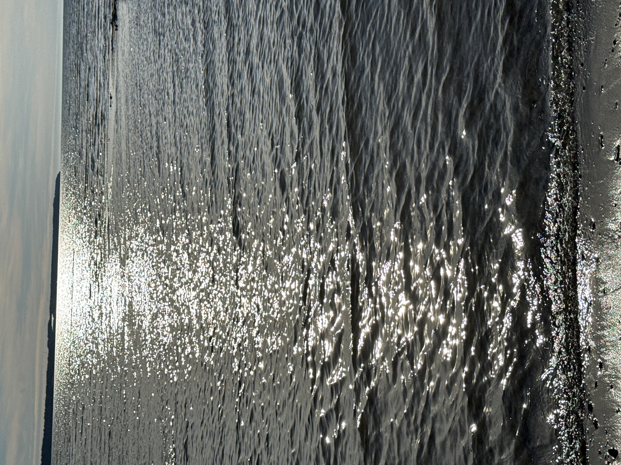 A sun-drenched, rippling body of water occupies the majority of the frame, reflecting the sky with a shimmering, broken pattern of light. In the distance, a dark, silhouetted shoreline stretches horizontally across the horizon, topped by a pale, overcast sky. The foreground shows a dark, wet beach or shore with small pebbles and debris scattered across it, also catching the light. The overall scene is one of a coastal environment under a bright, yet diffused, sky. This could be a view from one of Tallinn's many beaches, such as Pirita Beach, or from a promenade along the Baltic Sea coast within the city.