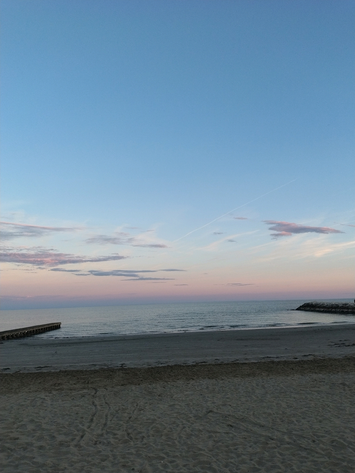 The image depicts a serene beach scene in Caorle, Italy, likely during twilight or early morning given the soft hues in the sky and on the water. The foreground is a sandy beach, showing subtle indentations and patterns from previous footsteps or the wind. Beyond the sand, the calm sea stretches towards the horizon. On the left side of the image, a wooden jetty or pier extends into the water, with its posts visible. On the right, a rocky breakwater juts out into the sea. The sky is a gradient of pale blue transitioning into soft pink and lavender tones, with thin wisps of clouds scattered across it, one of which has a faint contrail from an aircraft. The overall impression is one of tranquility and natural beauty. There are no people or animals visible in the frame.