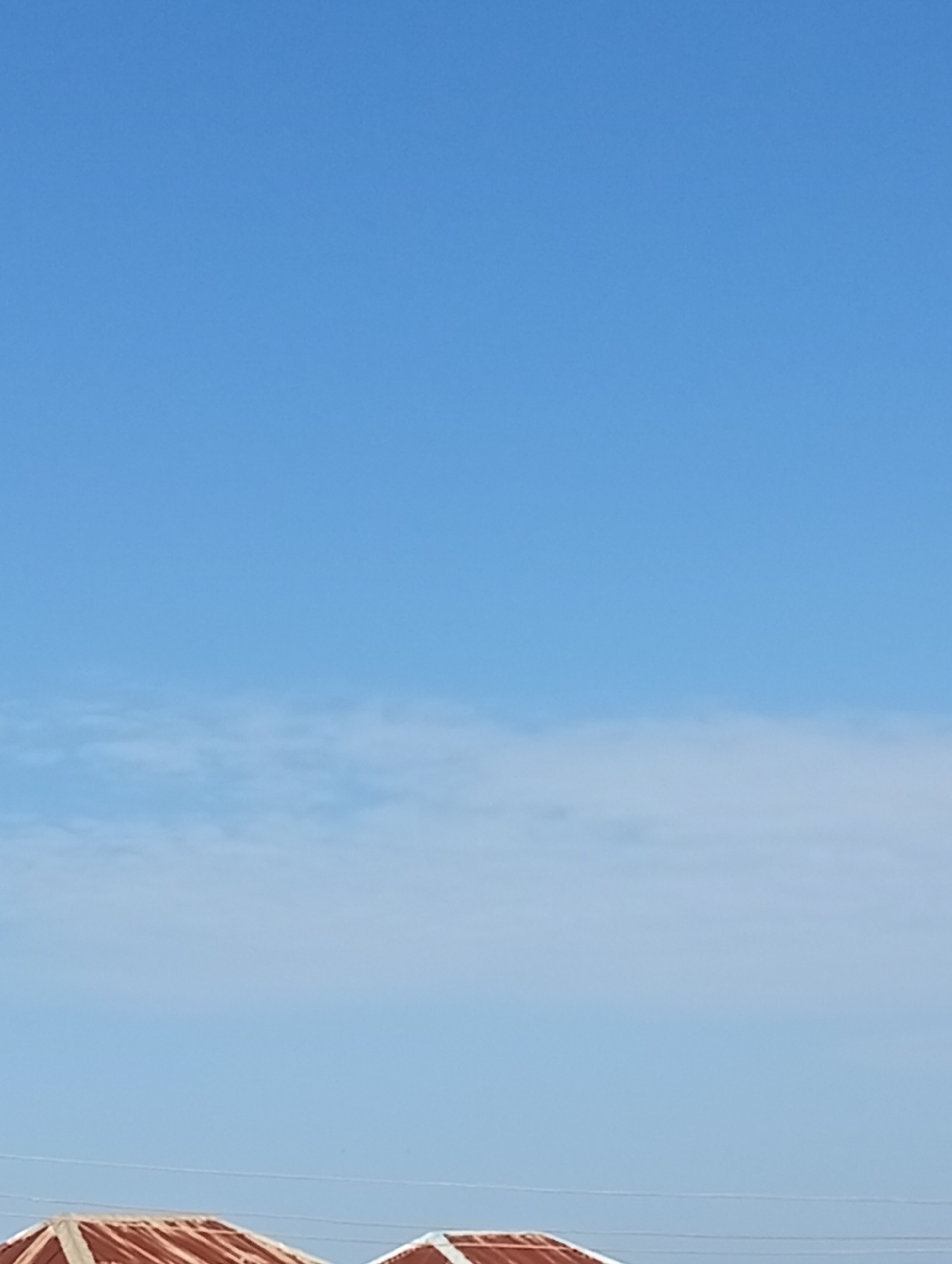 The image captures a section of the sky with faint wispy clouds. In the lower portion of the frame, the corrugated iron roofs of two buildings are visible. The roofs appear weathered, suggesting an outdoor setting in Bukuru, Nigeria. The sky is a clear, bright blue, indicating daylight. There are no people or discernible actions in the scene. The overall impression is one of a residential or utility area under a vast, open sky.