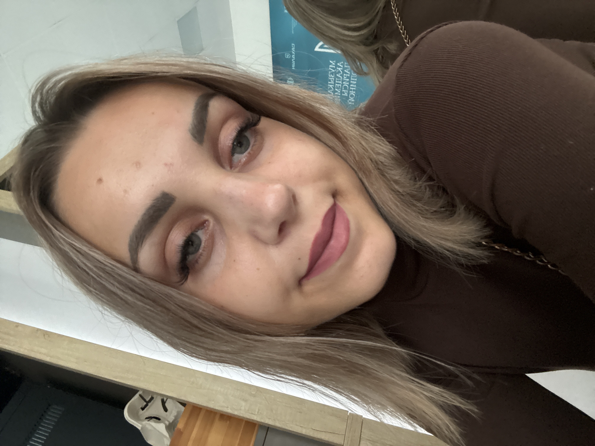 The image is a close-up, slightly angled selfie of a young Caucasian woman with blonde hair and light-colored eyes. She is wearing a dark brown, high-necked top. Her makeup includes noticeable false eyelashes, eyeshadow, and matte pink lipstick. She has freckles on her face and a mole on her forehead. The background is out of focus but appears to be an indoor setting with a light-colored wall and some objects that suggest a room, possibly with wooden elements. A turquoise banner with text in Russian is visible in the background, suggesting a public space. The woman's expression is neutral, with a slight smile. The image was taken in Yuzhno-Sakhalinsk, Russia.