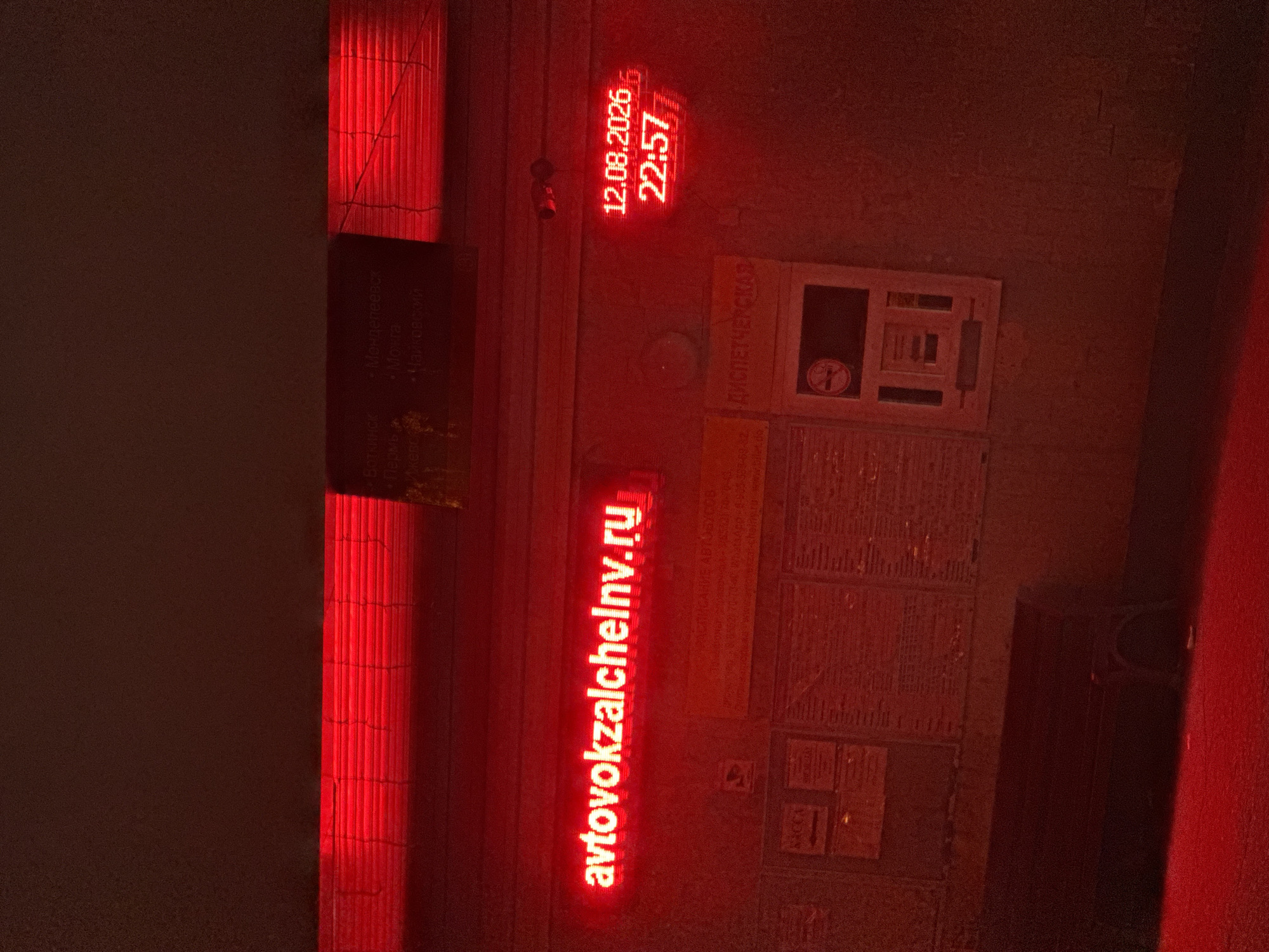 The image shows a nighttime scene at what appears to be a bus station. A prominent red neon sign displays the web address "avtovokzalchelny.ru". To the right, a digital clock shows the date "12.08.2026" and the time "22:57". In the background, there are bulletin boards with information and a vending machine or ticket dispenser. A bench is visible in the foreground. The overall lighting is red, suggesting the presence of red neon lights or a red filter over the camera lens. There are no people visible in the image. The scene is consistent with the exterior of a bus station in Oktyabr'-Bulyak, Russia, given the Russian text and the nature of the establishment.