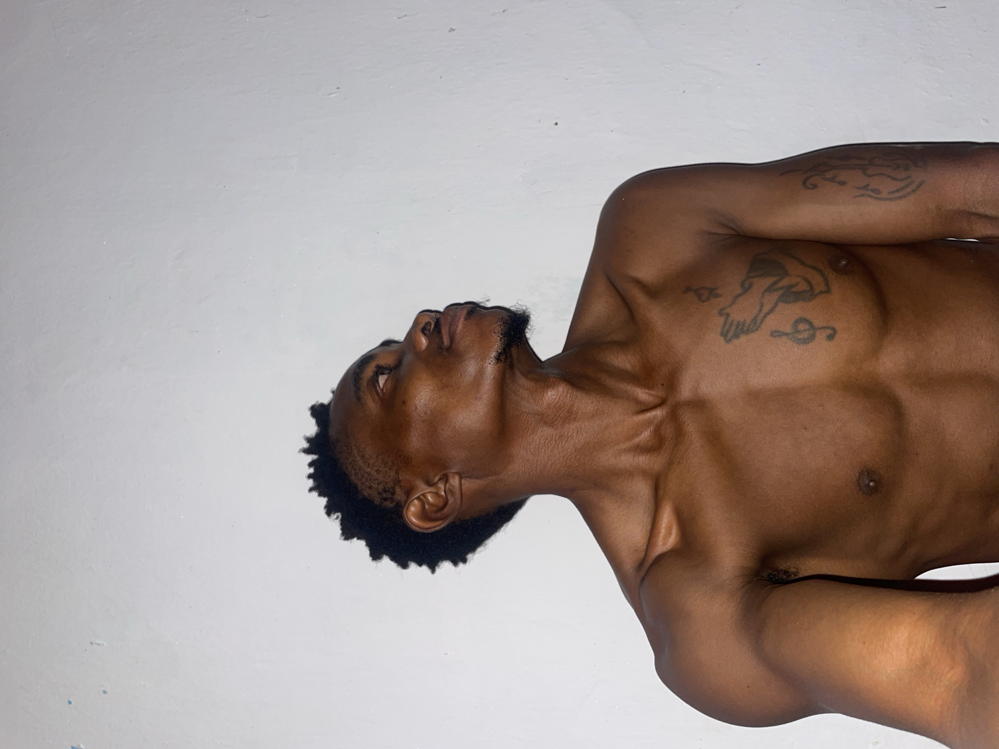A medium shot captures a shirtless Black man with dark skin and a short, textured hairstyle, looking up and to the right. He has a thin beard. Tattoos are visible on his chest and right arm: a pair of praying hands with musical notes on his chest, and an abstract design with musical notes on his arm. He stands against a white, textured wall. The lighting is dim, casting shadows on his face and body.