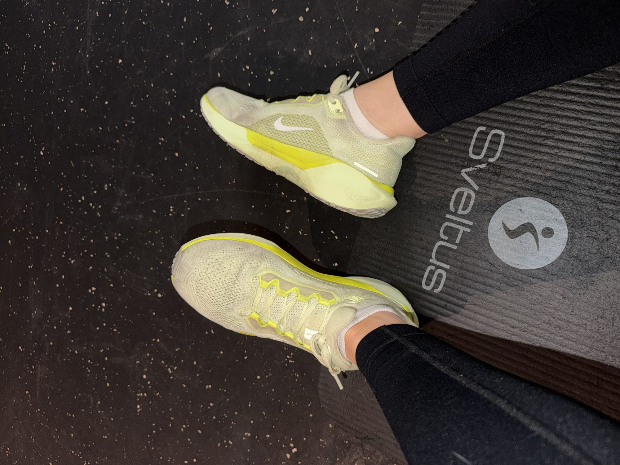 The image shows a first-person perspective of two feet wearing running shoes, presumably indoors. The shoes are pale yellow and white with a Nike swoosh on one. The person is wearing black leggings and white socks. The shoes rest on a dark surface, potentially a gym floor on the left and a yoga mat with the word "Sveltus" printed on it on the right. The location is unconfirmed as Paris, France.