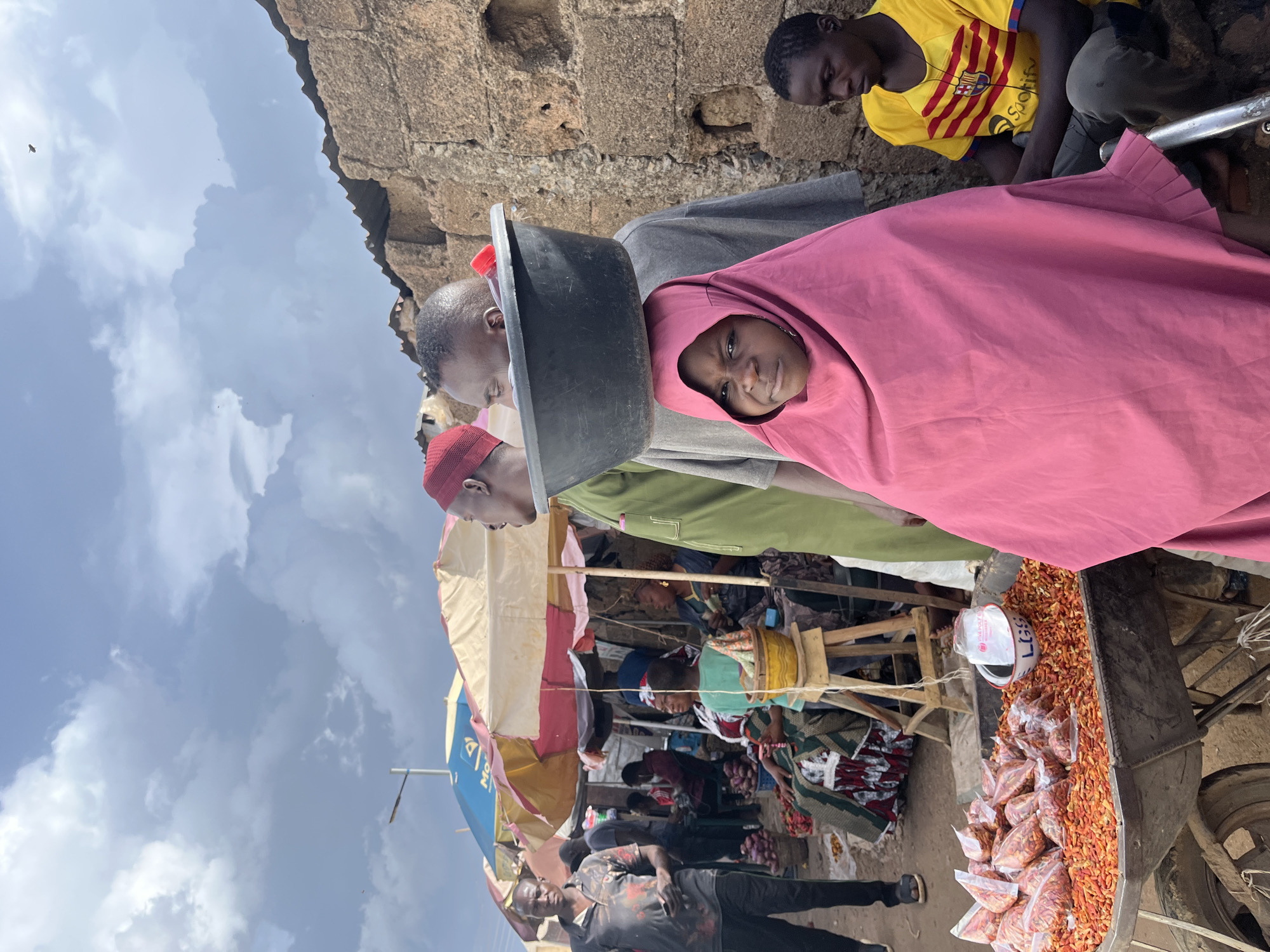 In Rafin Par, Nigeria, an outdoor market scene unfolds under a cloudy sky. A young girl, her face partially obscured by a vibrant pink hijab, stands in the foreground. Behind her, a man balances a large black basin on his head, his gaze directed to the left. To his right, a young man wearing a yellow and red striped t-shirt with a Spotify logo is seated. The market is populated with several individuals, some partially visible, navigating the stalls. Umbrellas provide shade for various vendors and their goods. To the left of the frame, a wheelbarrow overflows with small, bagged red peppers and loose orange chili peppers, with a white bowl placed amongst them. The background features a rustic stone wall.